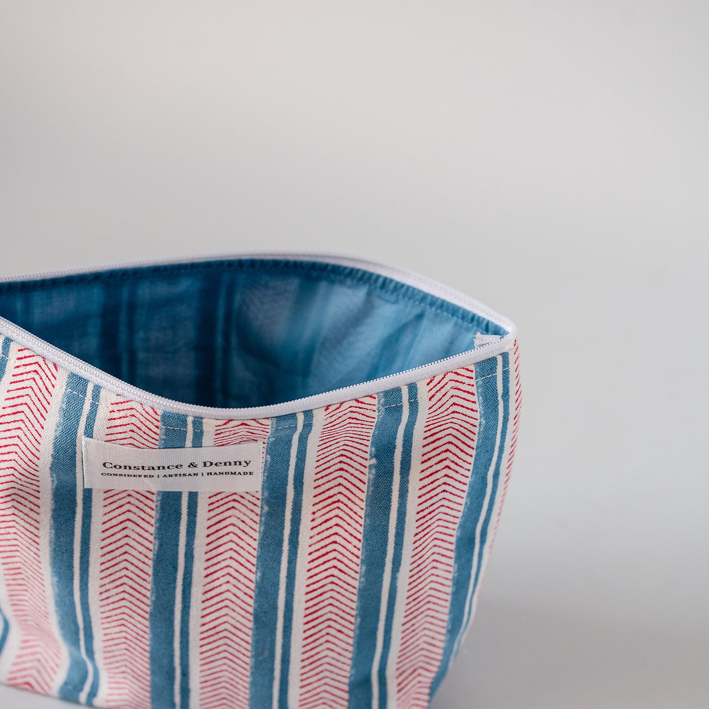 A close-up of a striped fabric cosmetic pouch with a blue denim-like interior lining. The exterior has vertical stripes in blue, red, and white, with a white label reading 'Constante & Deny' and 'Italian Leather & Furs'.