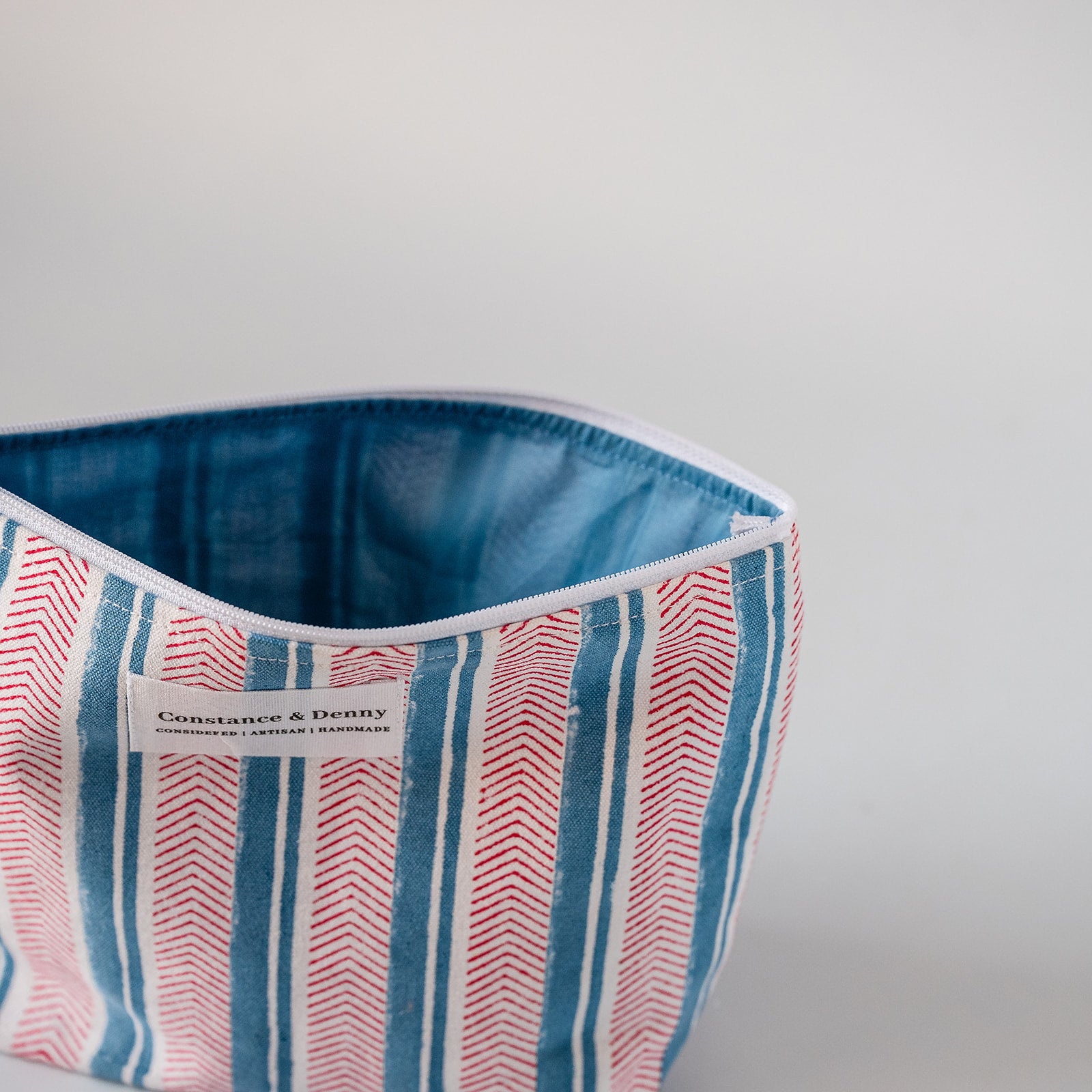 A close-up of a striped fabric cosmetic pouch with a blue denim-like interior lining. The exterior has vertical stripes in blue, red, and white, with a white label reading 'Constante & Deny' and 'Italian Leather & Furs'.