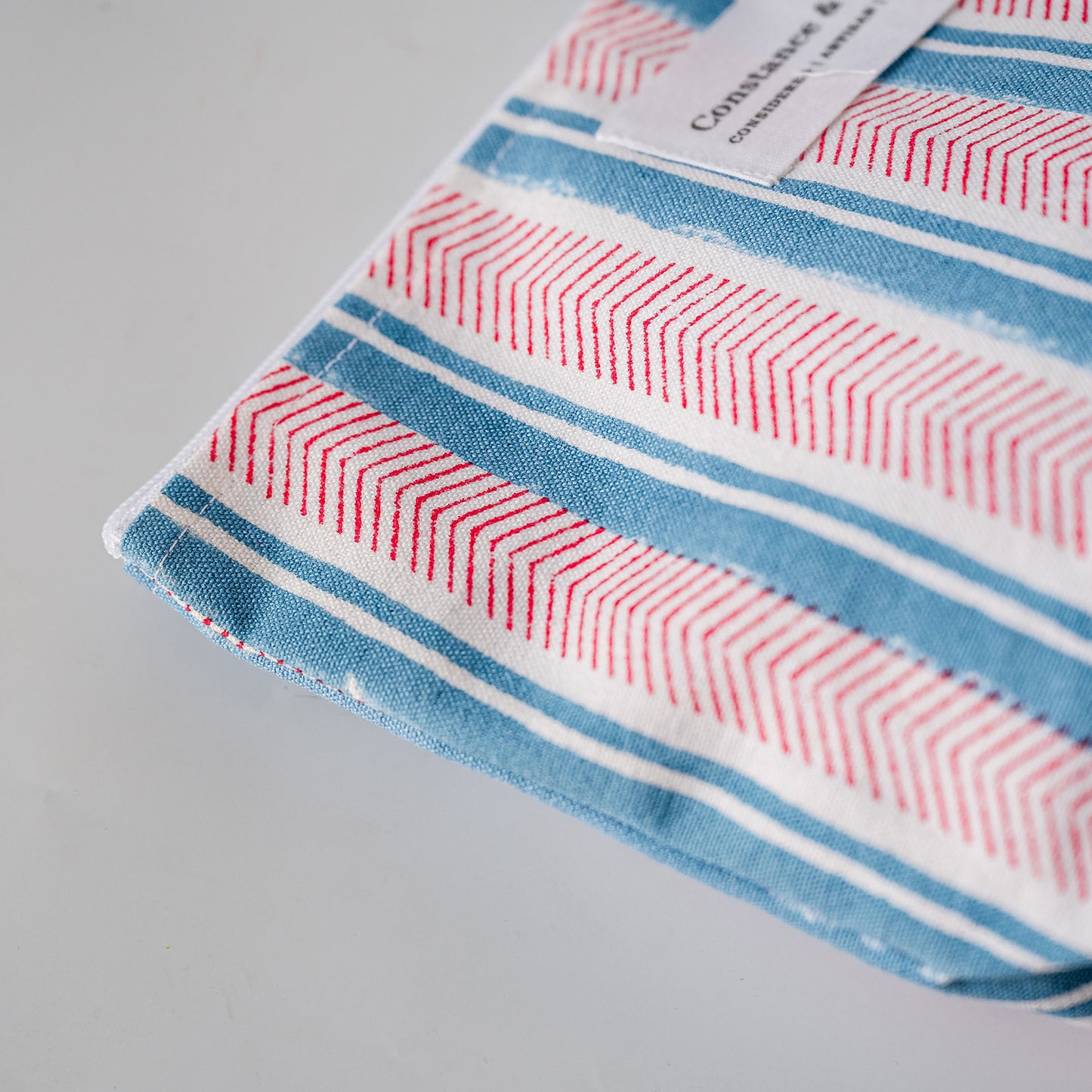 A close-up of a striped textile product, likely a towel or cloth, featuring blue, white, and red stripes with a red chevron pattern. A white paper label is visible near the top edge with the brand name 'Constable & Co'.