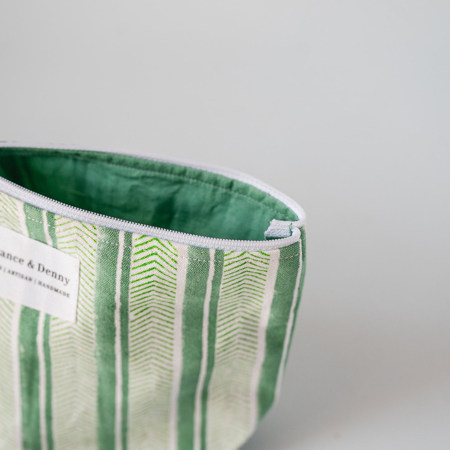 A close-up of a green and white striped fabric pouch with a white zipper and a white brand label that reads 'Dance & Denny'.