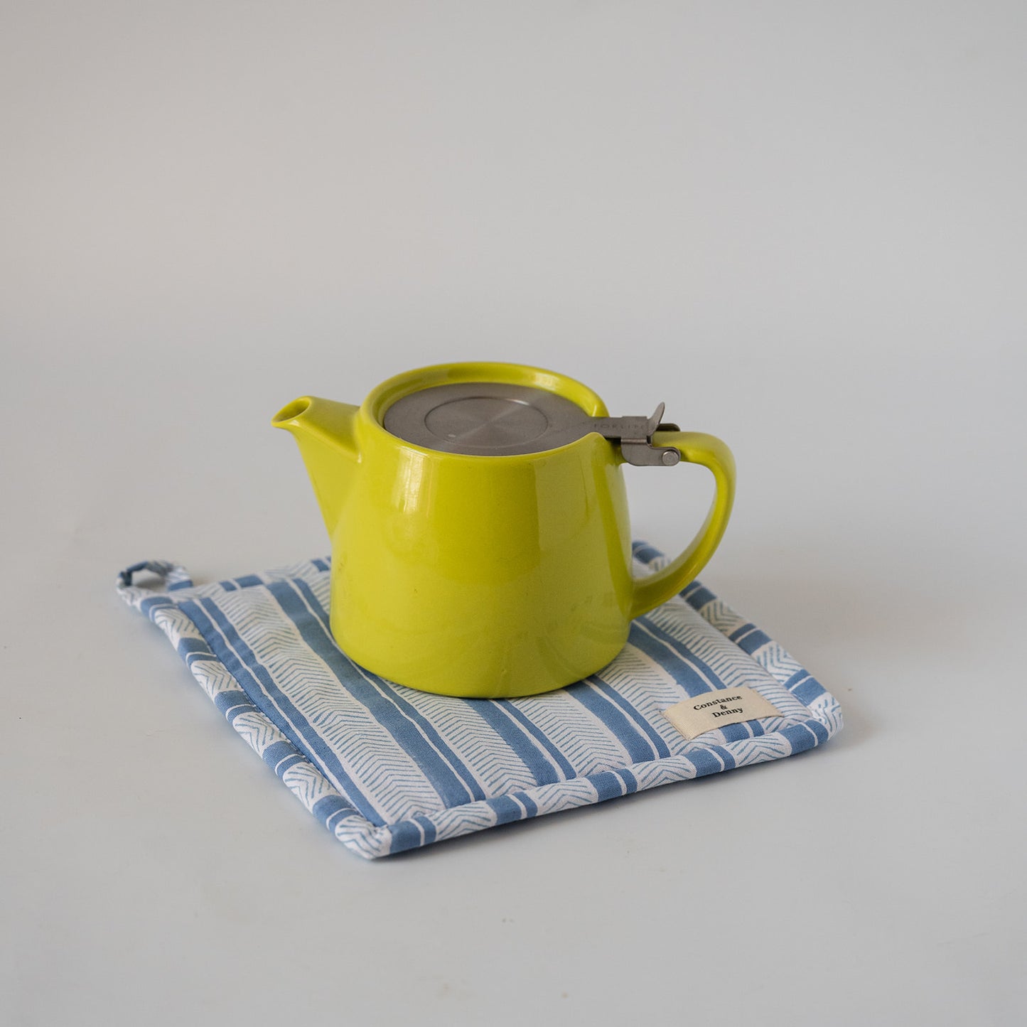 A bright yellow-green ceramic teapot with a stainless steel lid and handle clasp, resting on a blue and white patterned fabric pot holder against a light gray background.