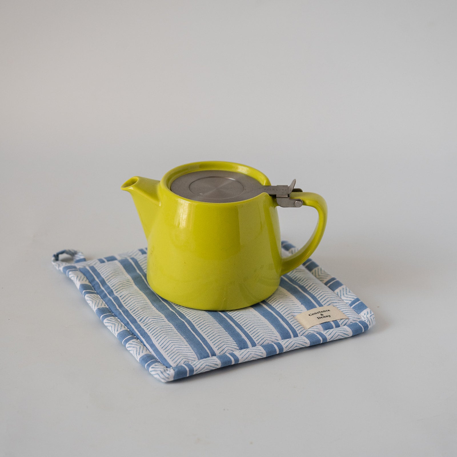 A bright yellow-green ceramic teapot with a stainless steel lid and handle clasp, resting on a blue and white patterned fabric pot holder against a light gray background.
