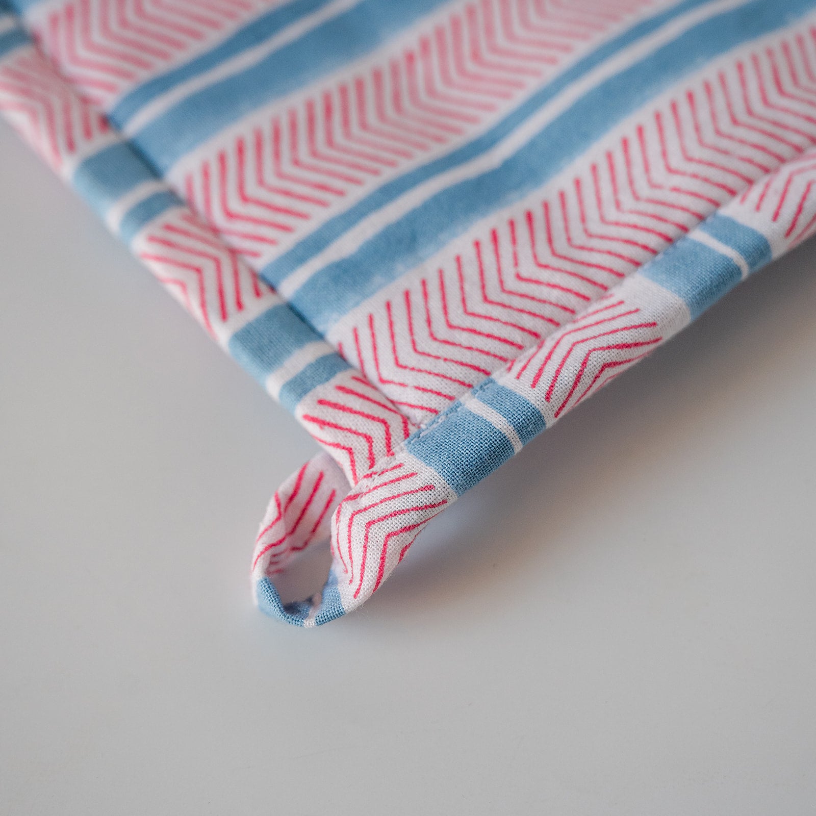 A close-up of a striped fabric product, likely a towel or cloth napkin, featuring red and white chevron patterns and light blue stripes, with a matching fabric loop tab.