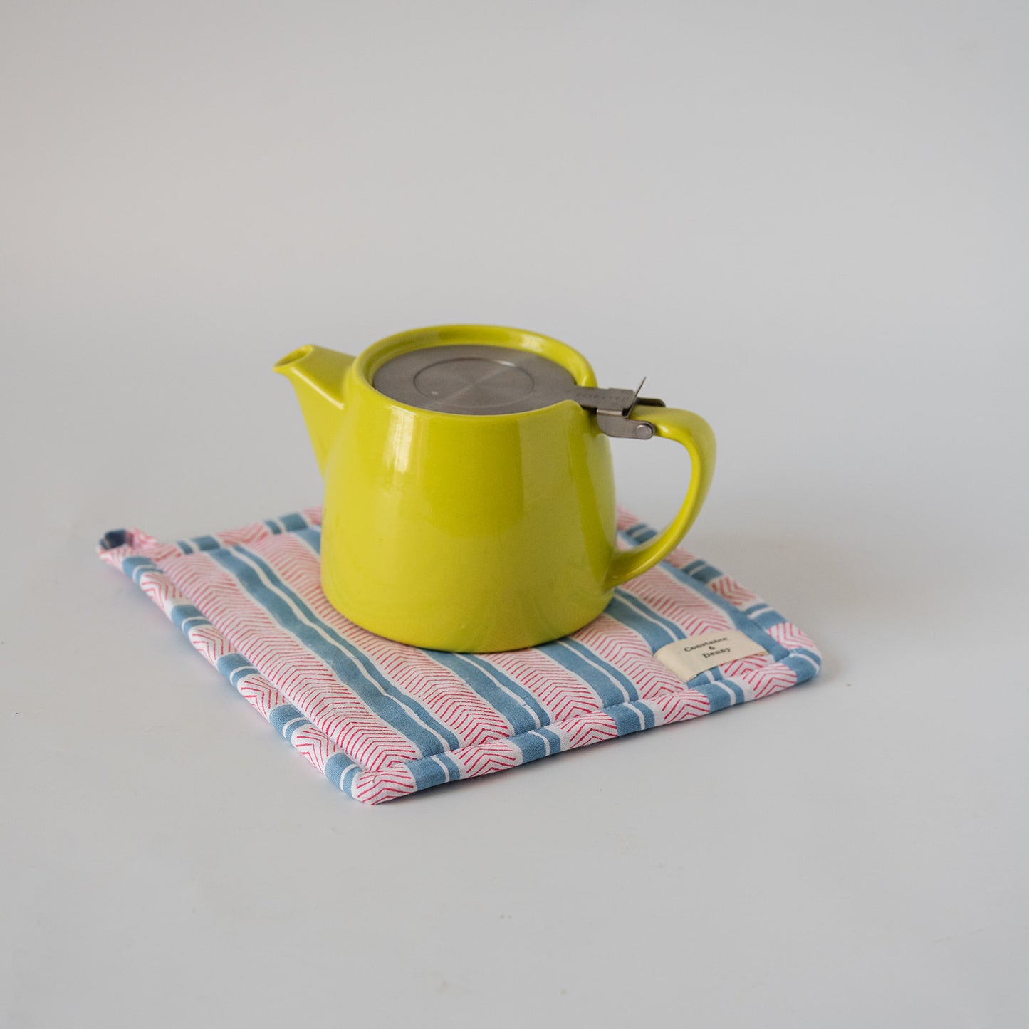 A bright yellow-green ceramic teapot with a stainless steel lid sits on a pink, blue, and white striped fabric tea towel against a plain light gray background.