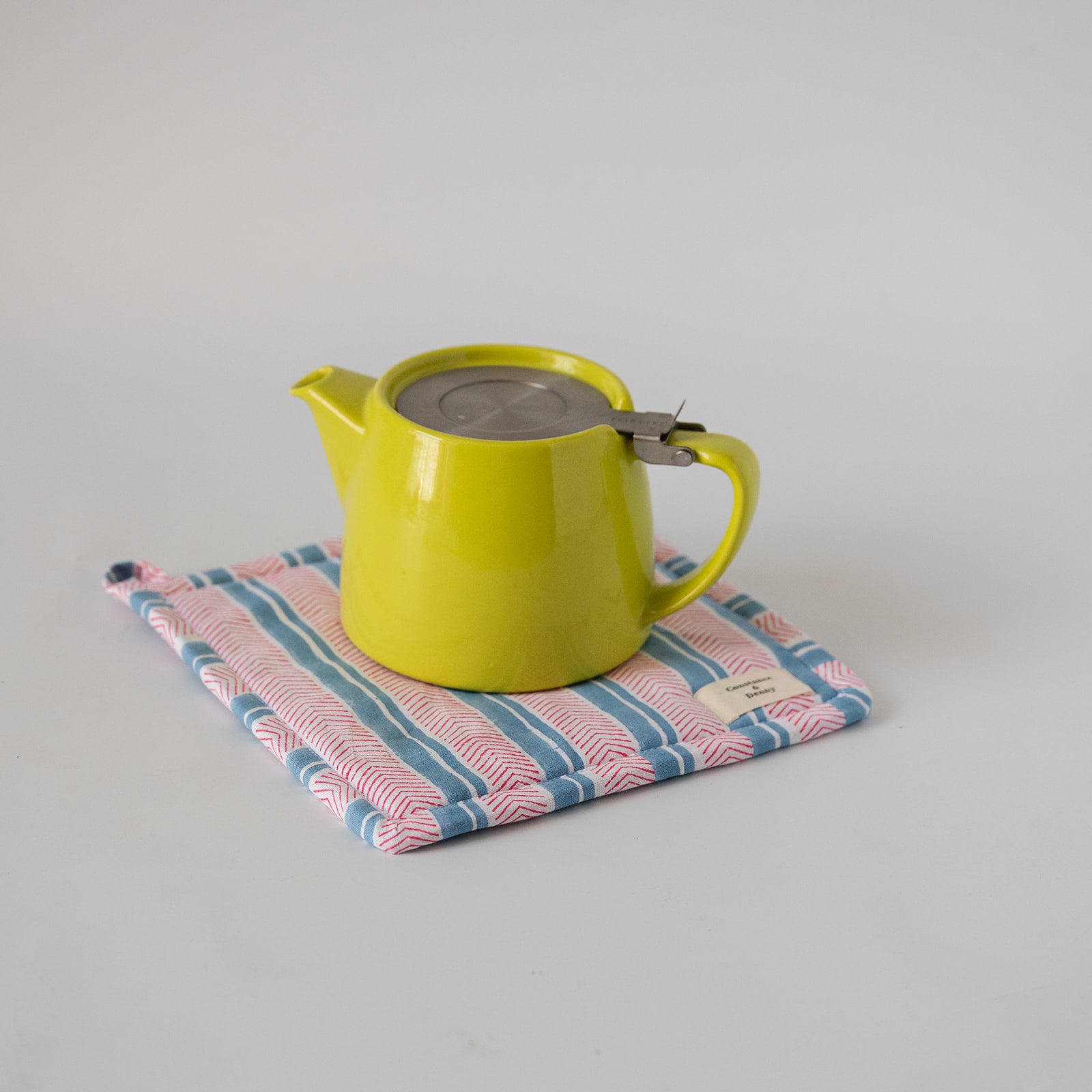 A bright yellow-green ceramic teapot with a stainless steel lid sits on a pink, blue, and white striped fabric tea towel against a plain light gray background.