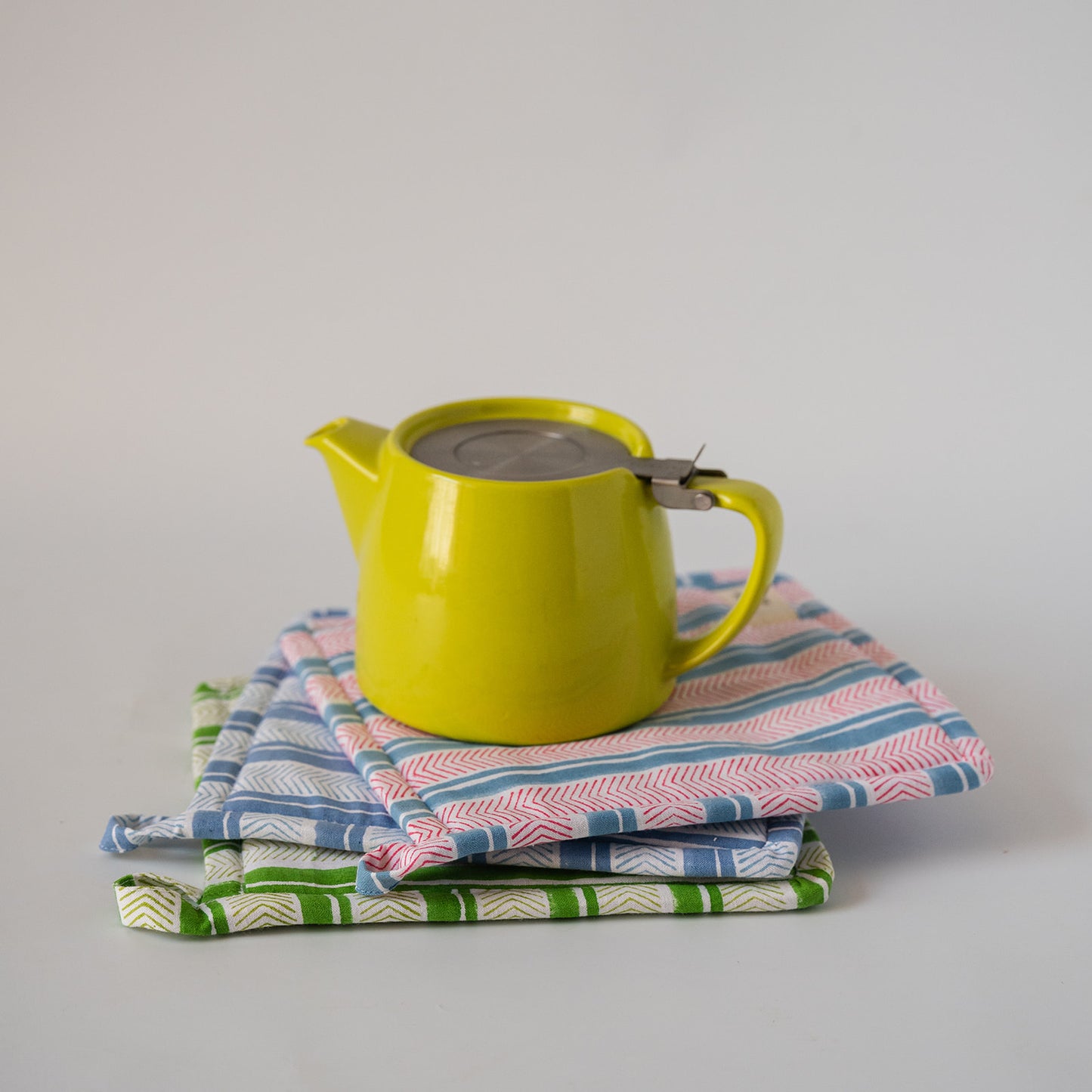 A bright yellow teapot with a metal lid sits on top of a stack of patterned fabric potholders on a light gray background.