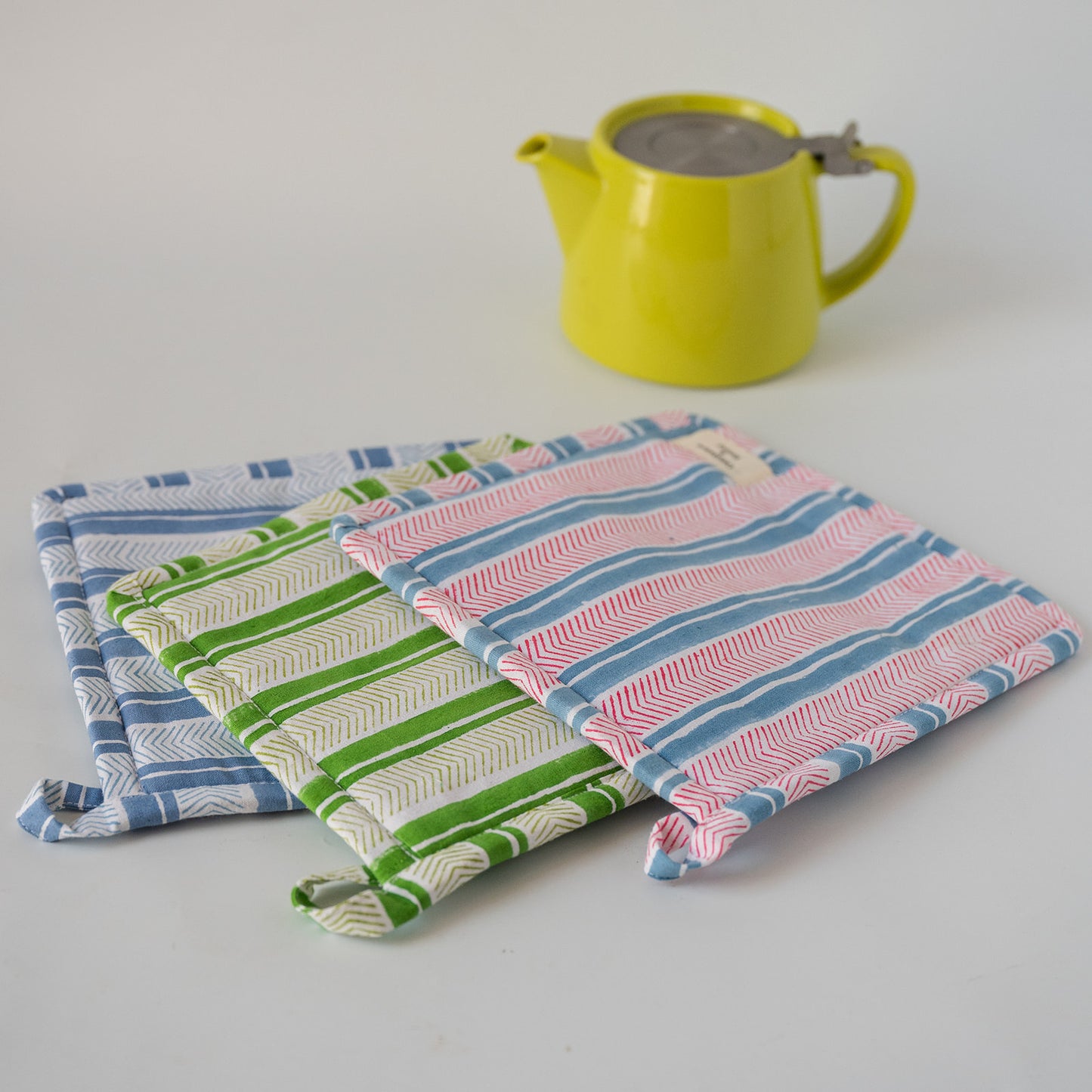 Three striped fabric potholders in blue, green, and pink are arranged on a white surface, with a bright yellow teapot in the background.