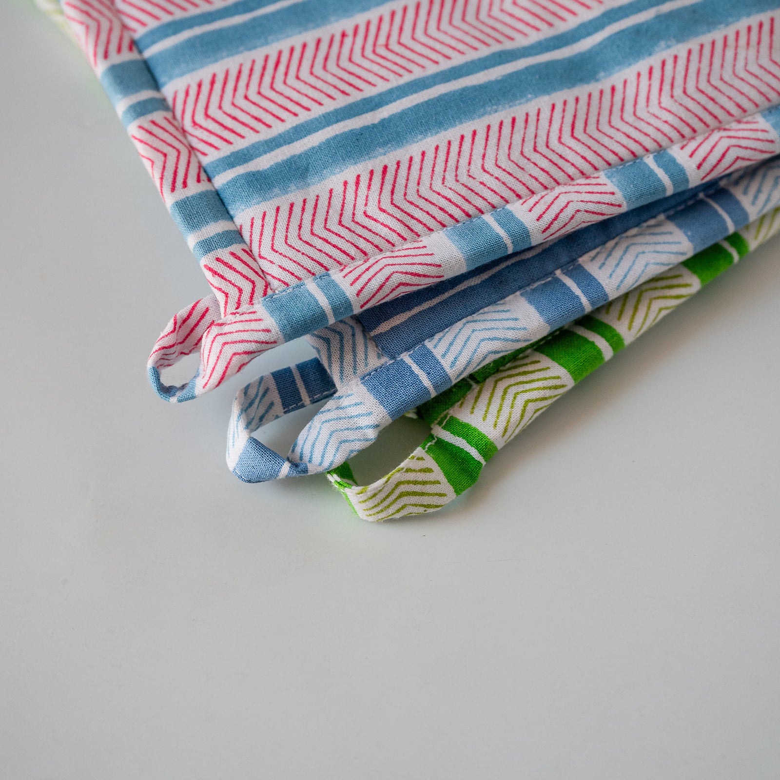 Three folded fabric potholders with colorful geometric patterns, including stripes, chevrons, and dots, arranged on a light gray surface.