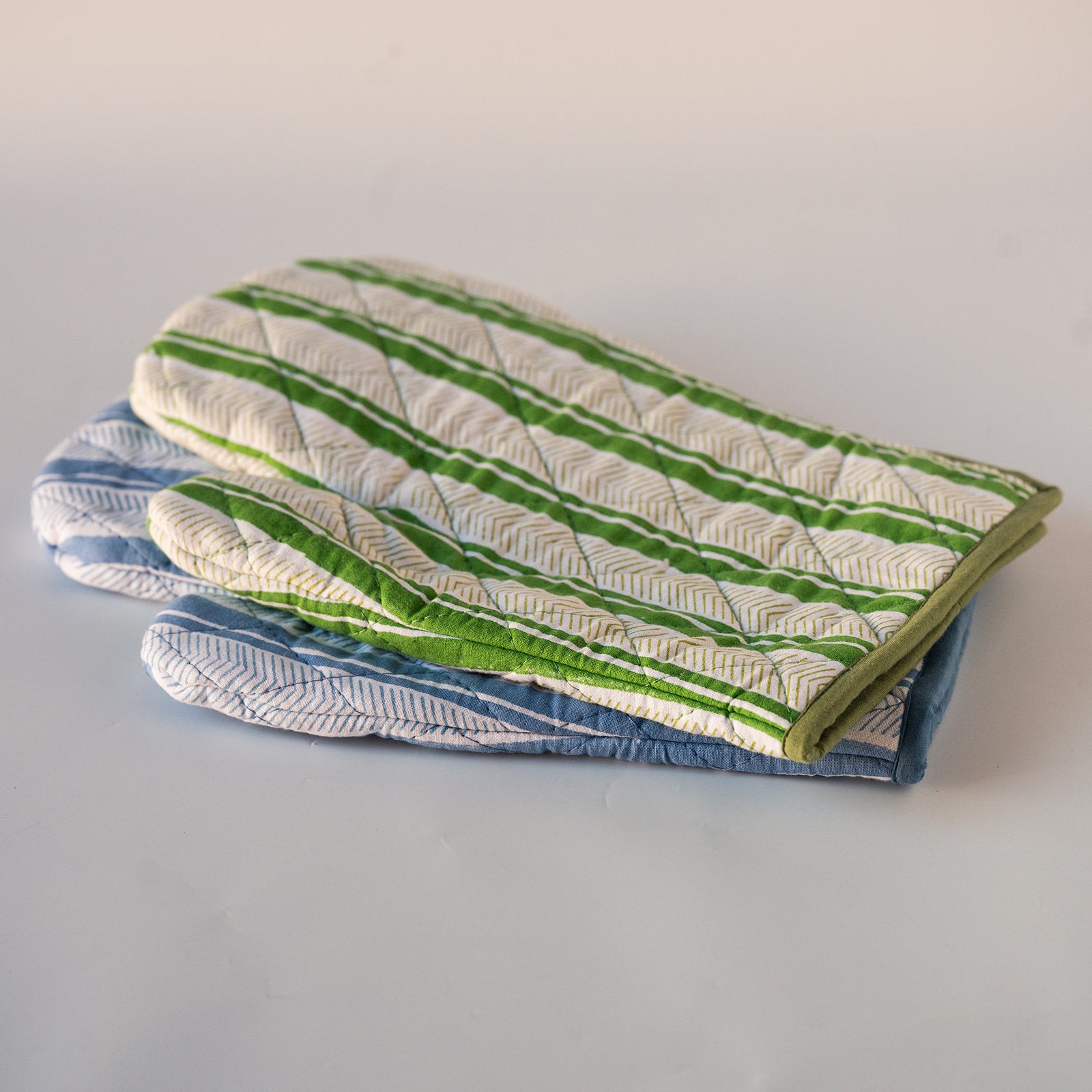 Two quilted fabric oven mitts stacked on a light gray background. The top mitt has a white base with green and beige herringbone stripes, and the bottom mitt has a blue and white herringbone pattern.