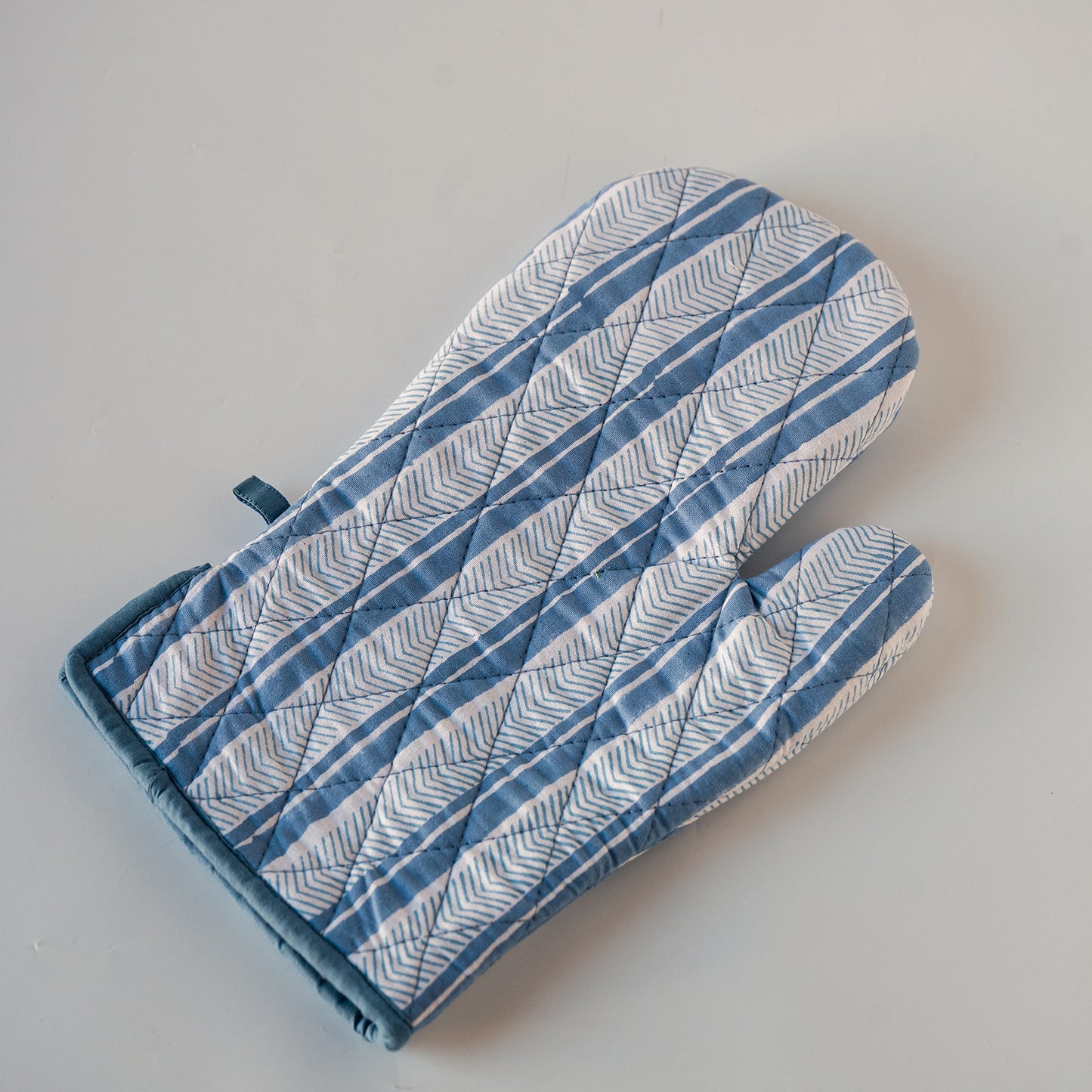 A quilted oven mitt with a blue and white herringbone pattern and solid blue trim, shown on a neutral light gray background.