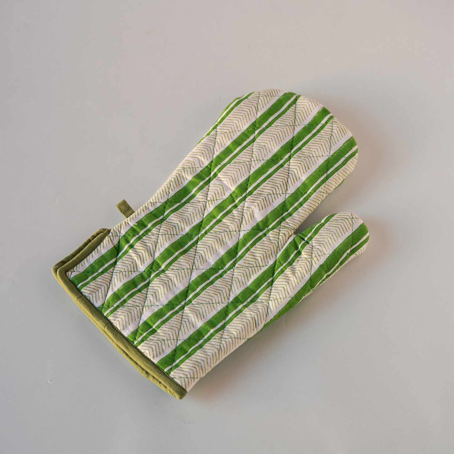 A single quilted oven mitt with a green and white striped pattern and olive green trim, photographed on a neutral light gray background.