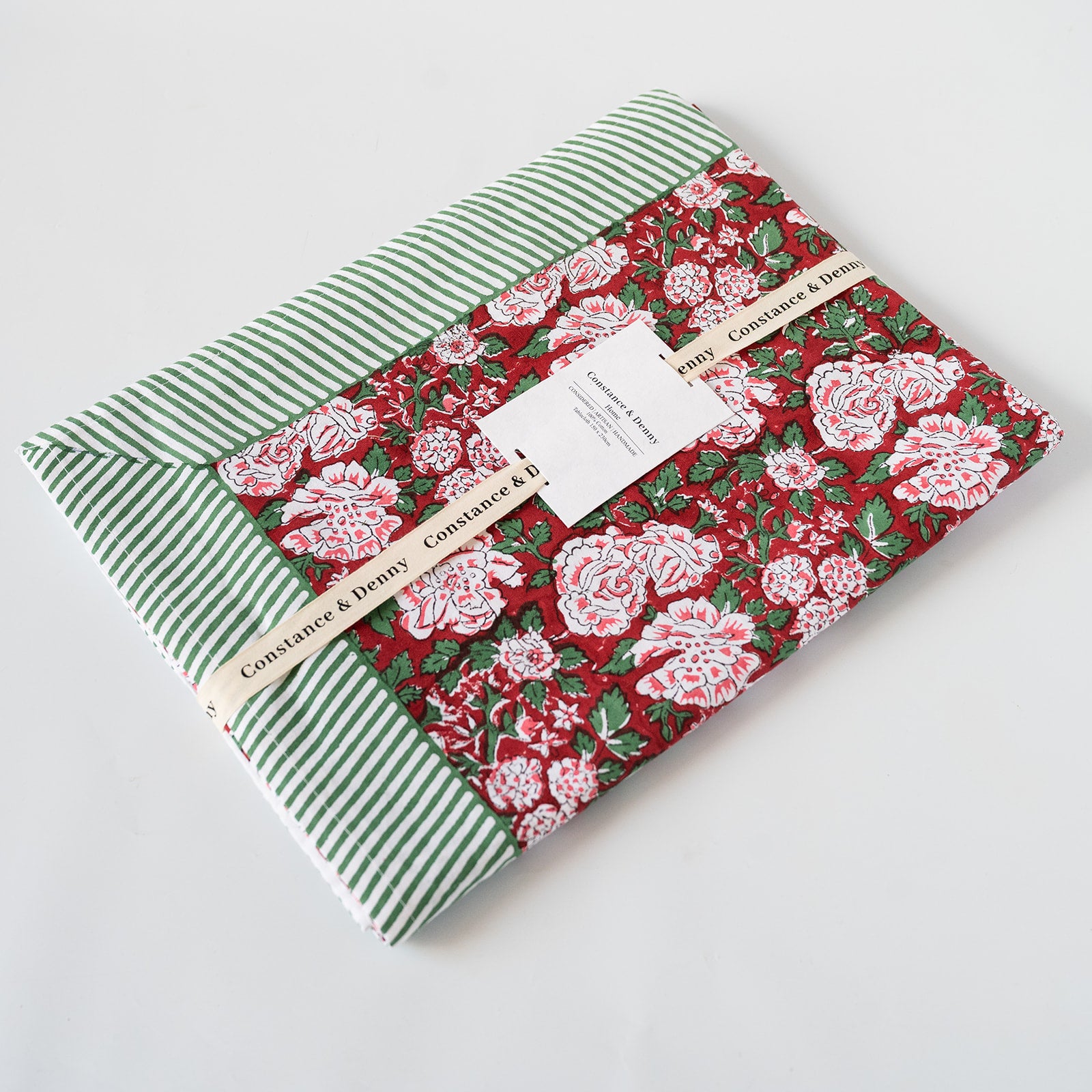 A folded fabric item, likely a quilt or blanket, showing three distinct patterned sections: a green and white striped section, a red floral section with pink and white flowers, and a red striped section. A paper label from 'Constance & Henry' is wrapped around the item, photographed on a clean white background.