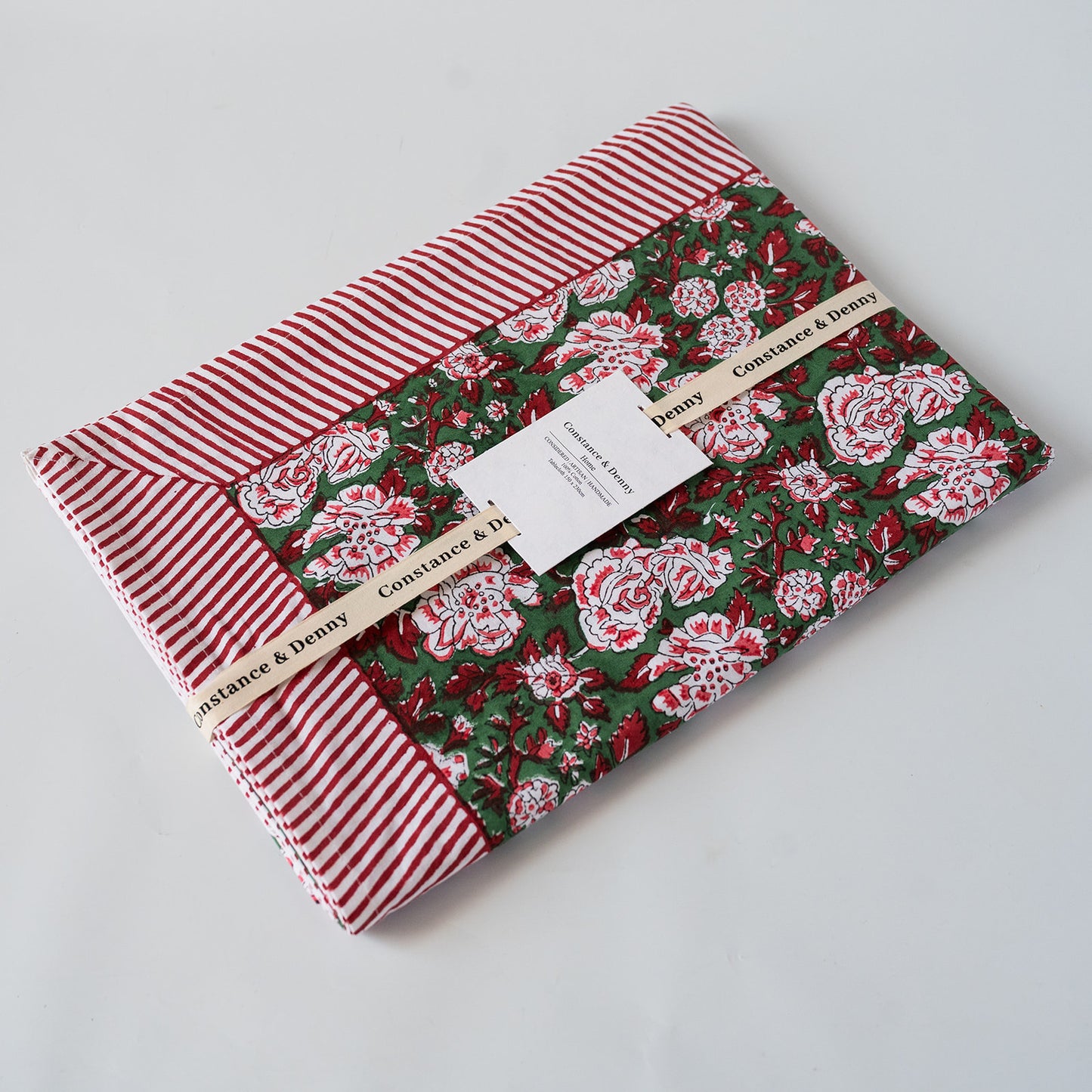A folded fabric item, likely a quilt or blanket, featuring a red and white striped border and a central green floral pattern with red and white roses. A paper tag and ribbon label reading 'Constance & Deny' are attached, photographed on a clean white background.