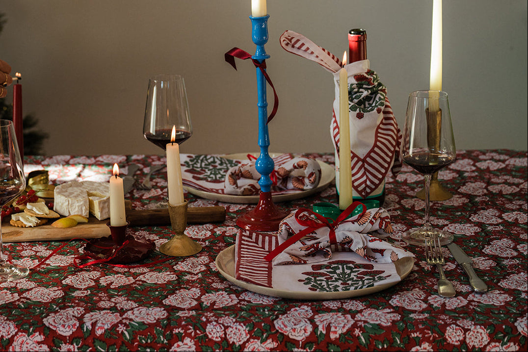 A festive holiday table setting with a red and white patterned tablecloth, lit taper candles in blue and gold holders, wine glasses, cheese on a board, and a person's arm visible on the left adjusting a candle. A decorated Christmas tree stands in the background.