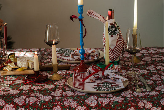 A festive holiday table setting with a red and white patterned tablecloth, lit taper candles in blue and gold holders, wine glasses, cheese on a board, and a person's arm visible on the left adjusting a candle. A decorated Christmas tree stands in the background.