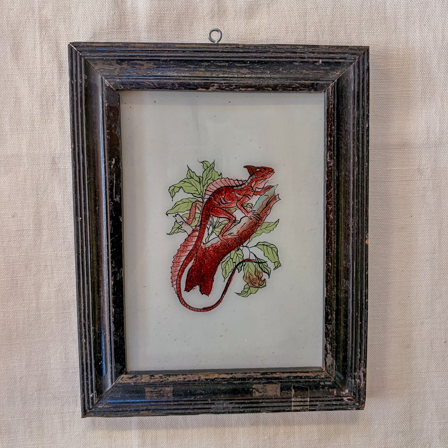 Embroidered red lizard on a white background in a dark wooden frame.