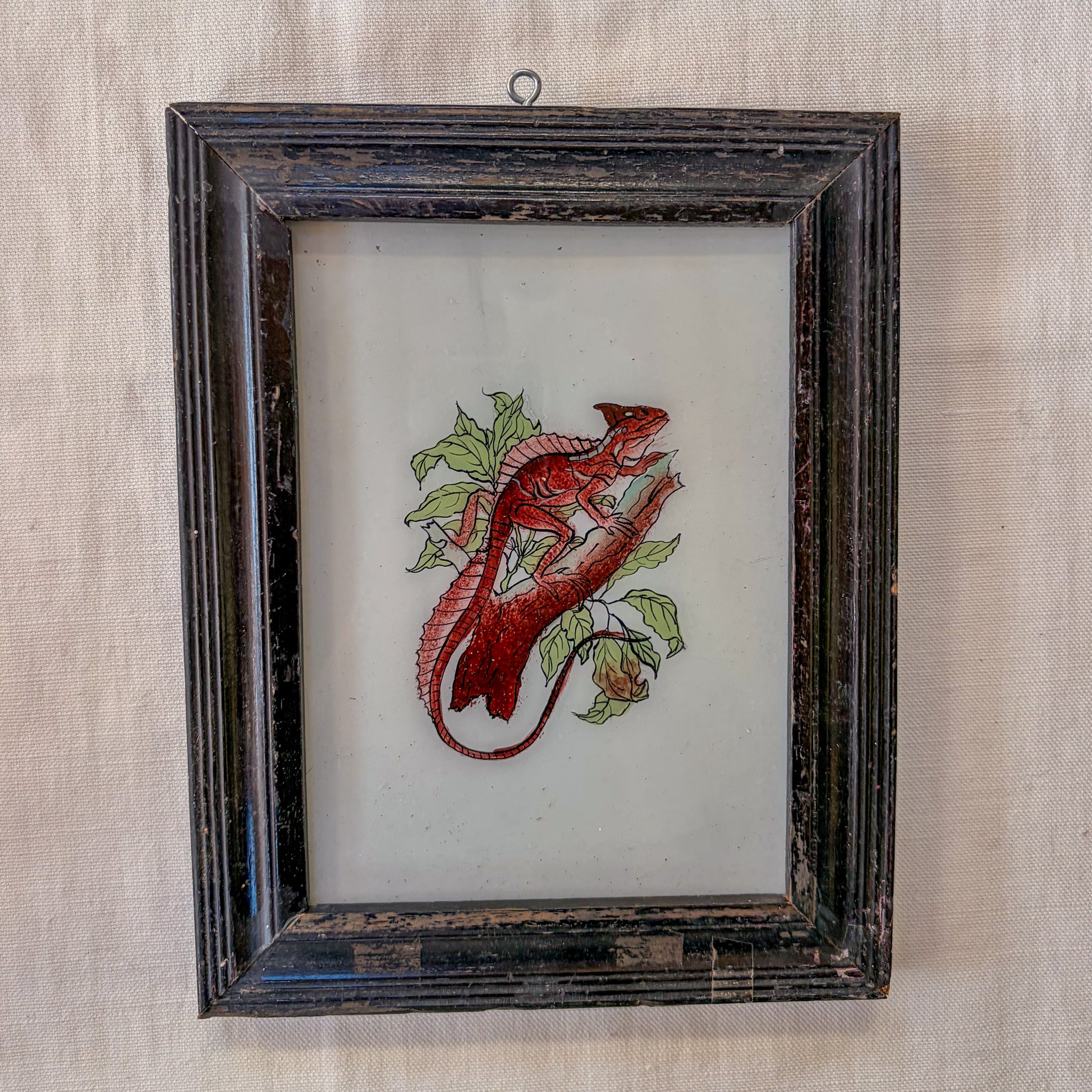 Embroidered red lizard on a white background in a dark wooden frame.