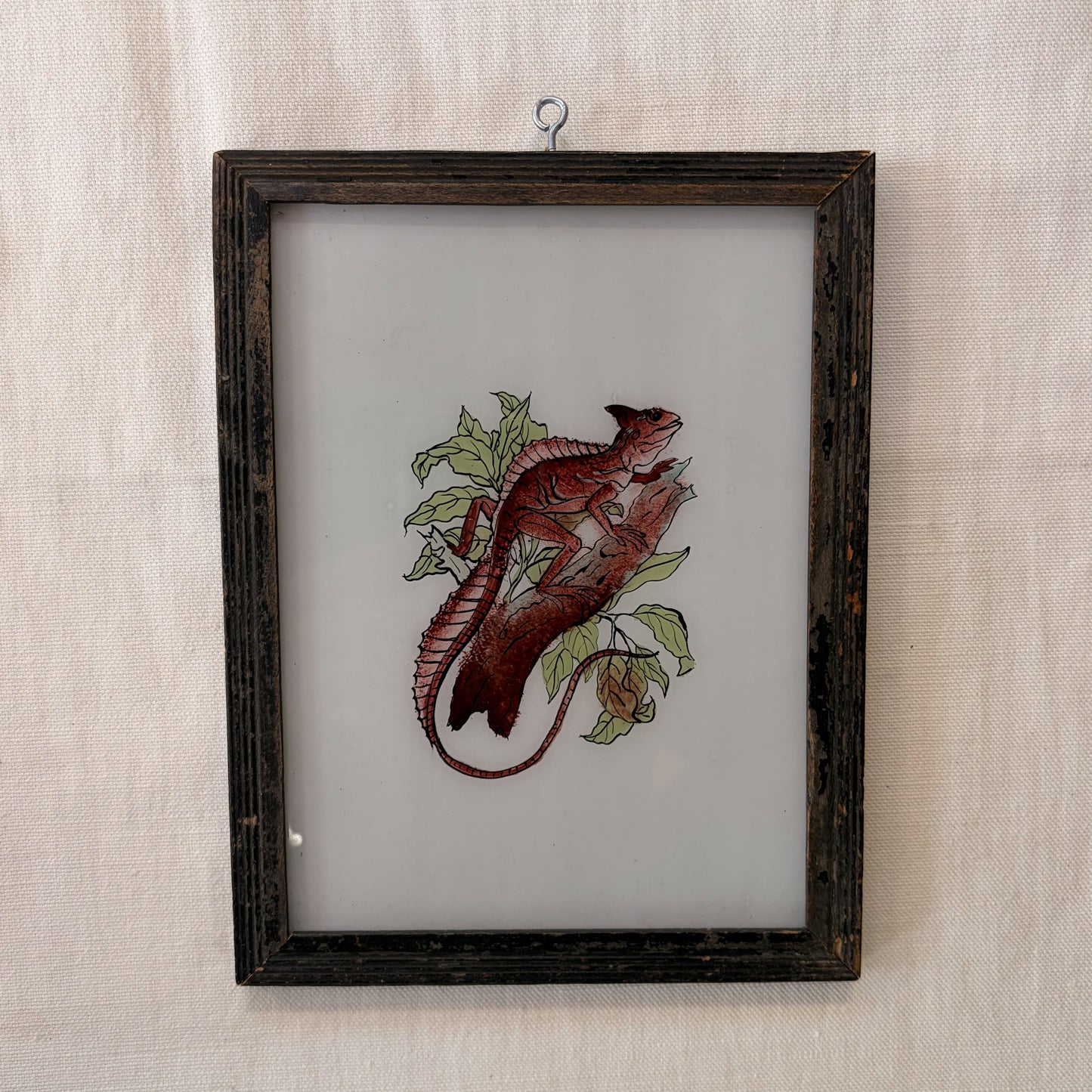 Embroidered lizard on fabric in a wooden frame against a textured beige wall.