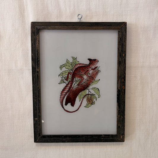 Embroidered lizard on fabric in a wooden frame against a textured beige wall.