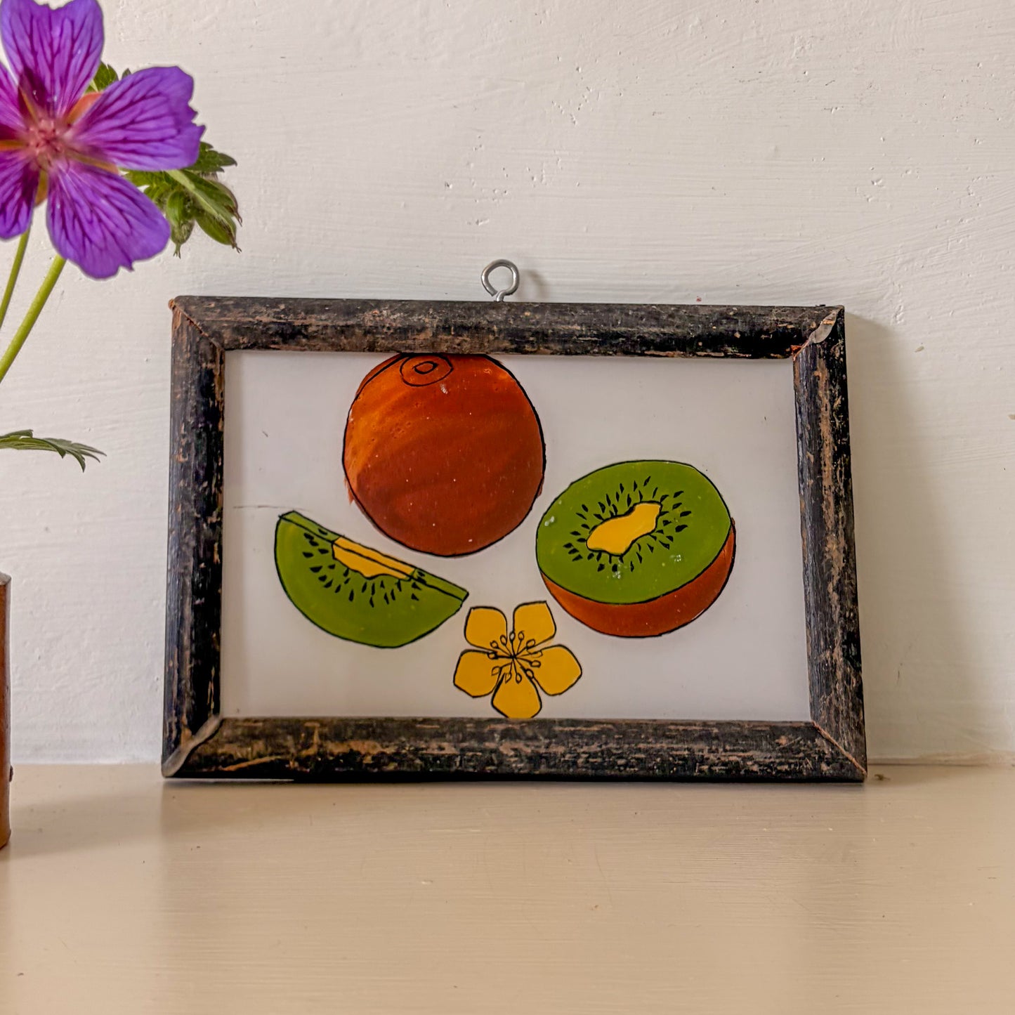 Framed artwork of fruits and a flower on a white wall with a purple flower in the corner.