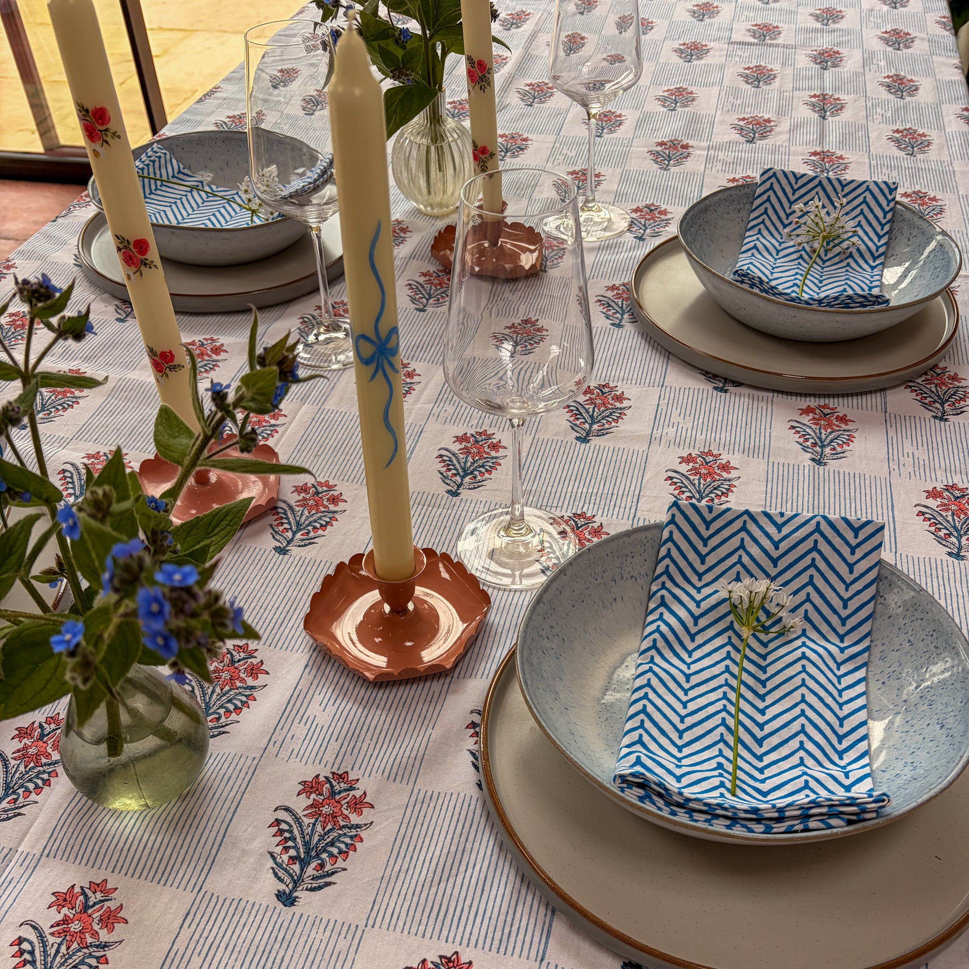 Dining table setting with floral tablecloth, plates, glasses, and decorative candles.