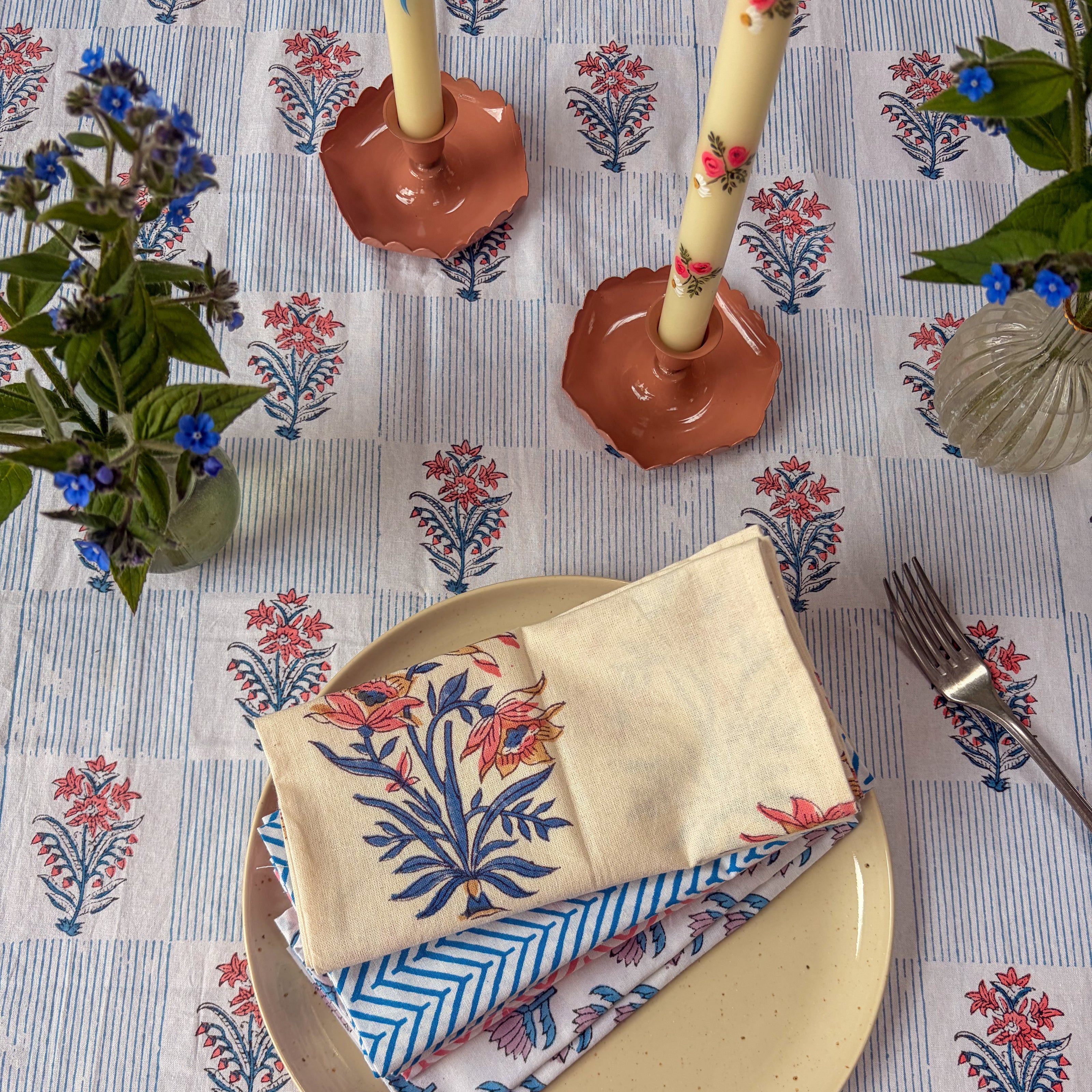 Decorative table setting with floral-patterned napkins, candles, and cutlery on a matching tablecloth.