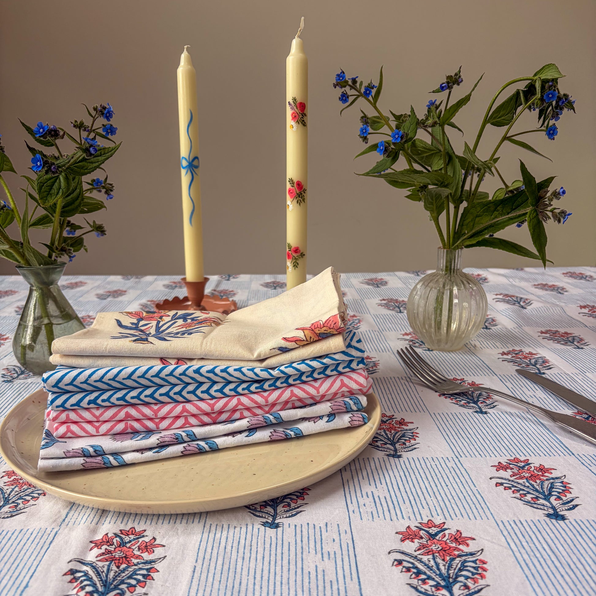 Stack of patterned napkins on a plate with candles and flowers on a tablecloth.