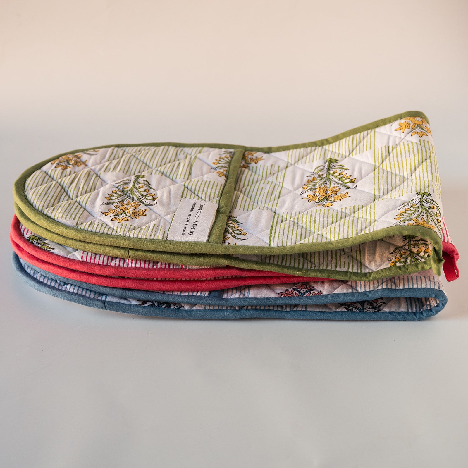A folded quilted fabric oven mitt or potholder with a floral and striped patchwork design, olive green trim, and a red handle, photographed on a plain light background.