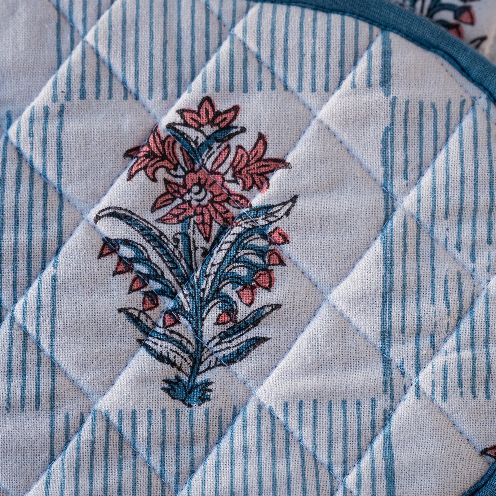Close-up of a quilted fabric item, likely a pot holder or coaster, featuring a white background with light blue vertical stripes, a diamond-quilted pattern, and a central floral motif in pink and blue.