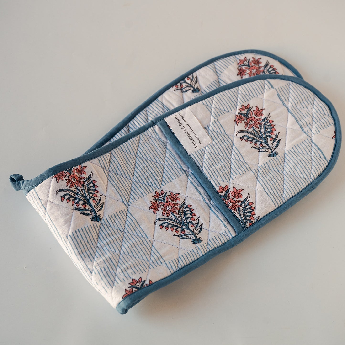 Two quilted cotton oven mitts with a blue and white striped and floral block print pattern, featuring a solid blue trim and a white brand label.