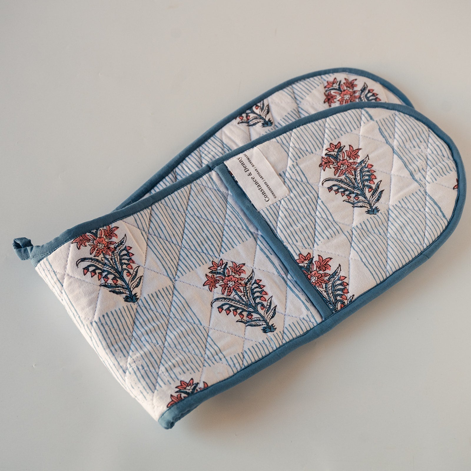 Two quilted cotton oven mitts with a blue and white striped and floral block print pattern, featuring a solid blue trim and a white brand label.
