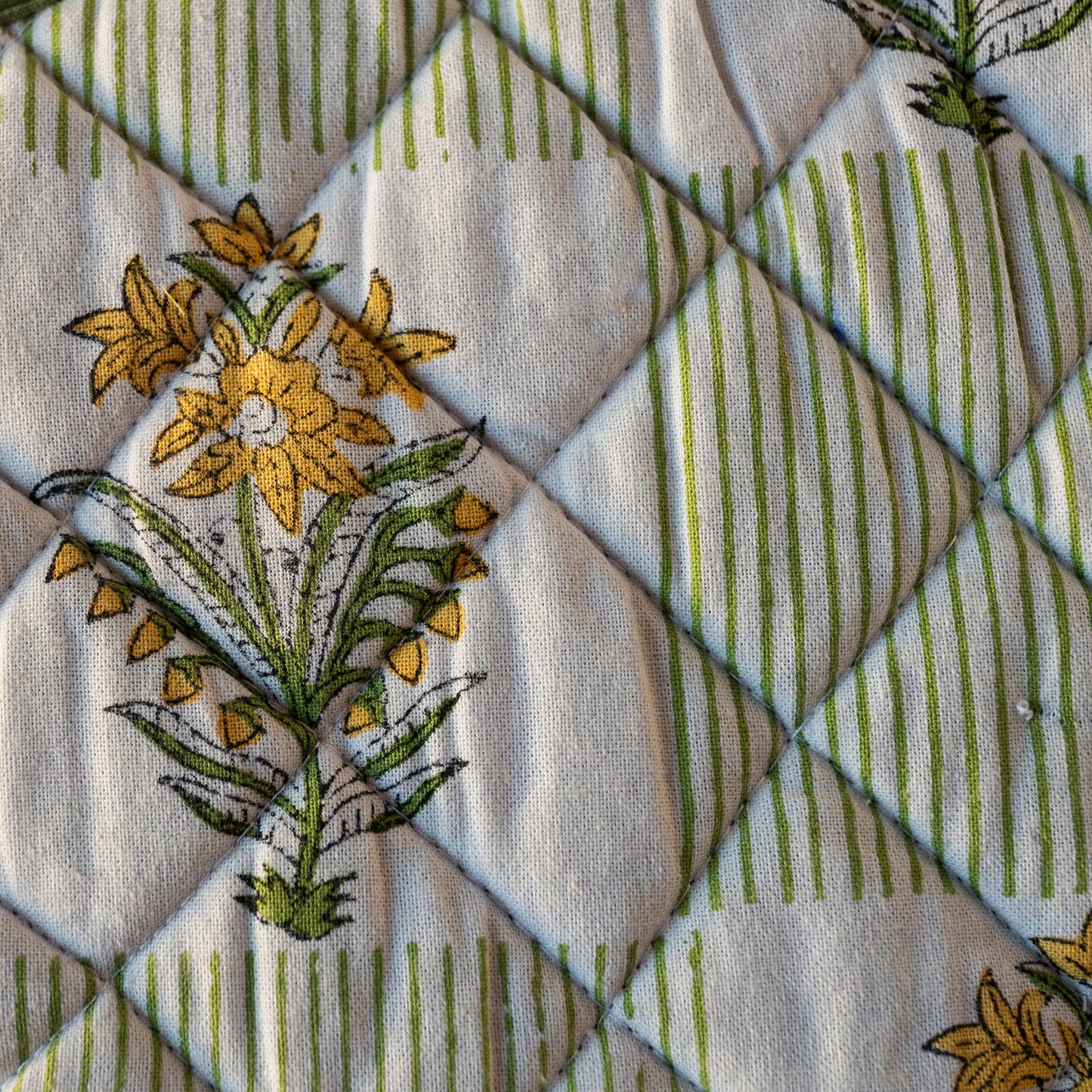 Close-up of a quilted fabric with a diamond stitched pattern, featuring yellow floral motifs and green vertical stripes on an off-white background.