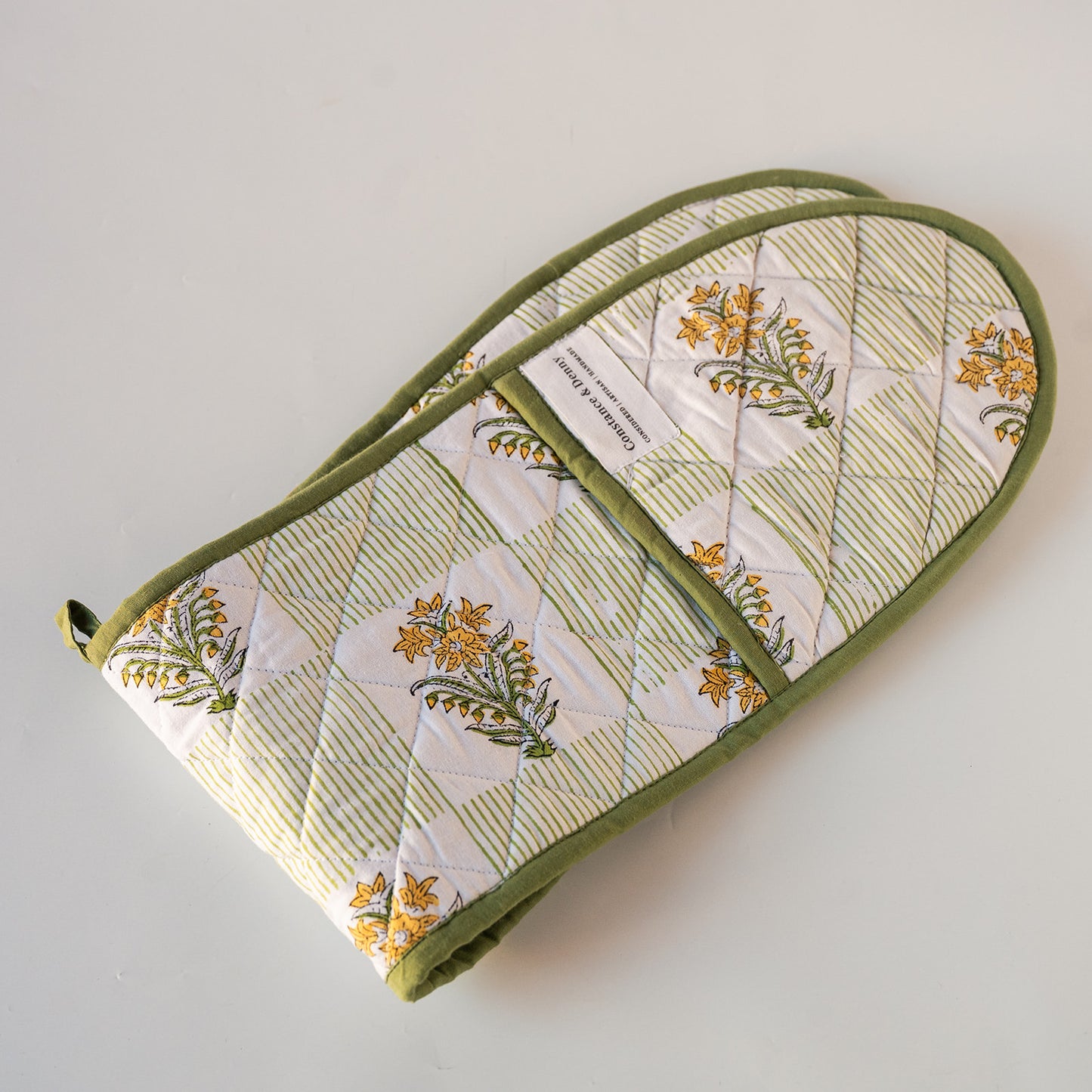 A quilted fabric oven mitt with a green border and a floral print on a white background, featuring a diagonal zippered pocket. The mitt is photographed on a plain, light off-white surface.