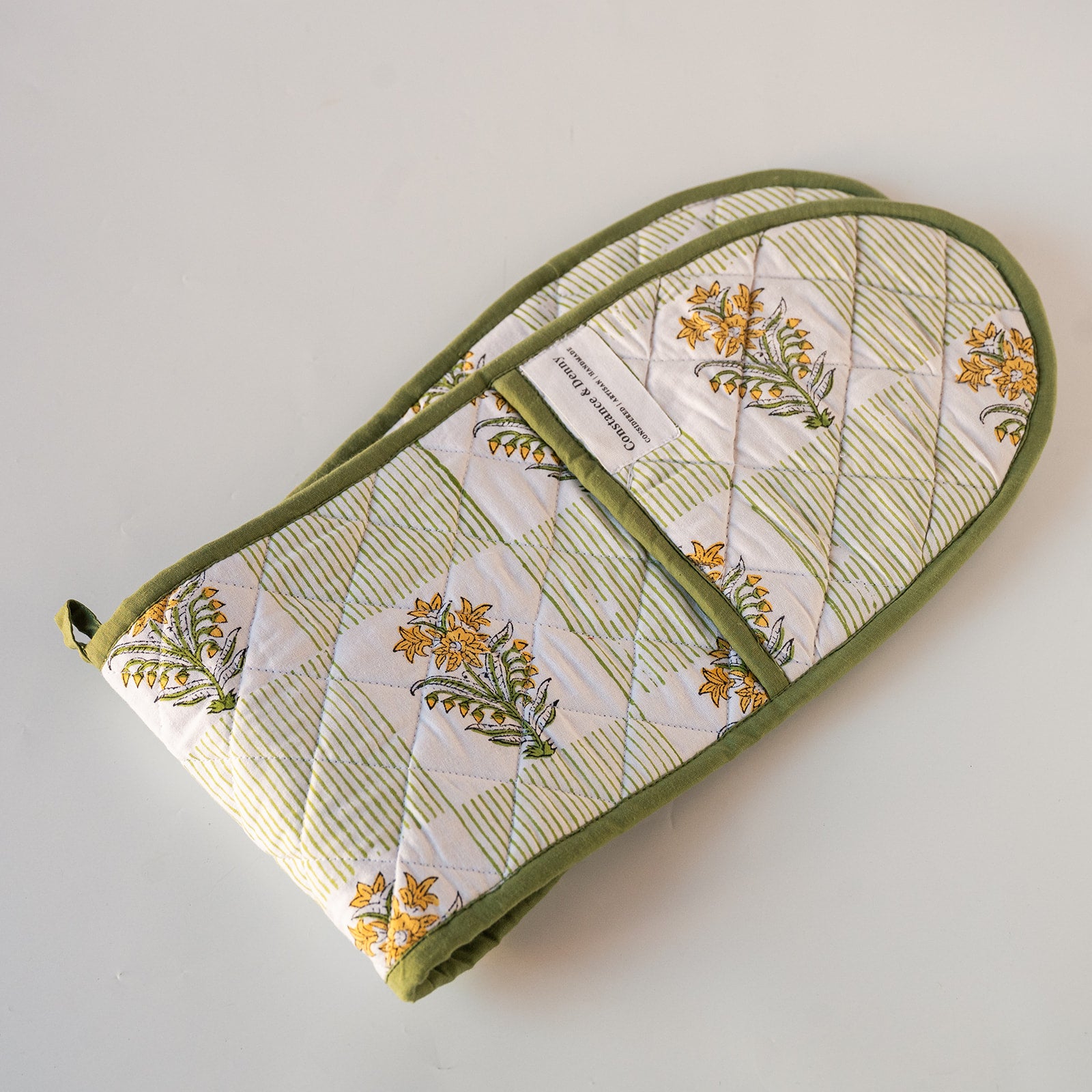 A quilted fabric oven mitt with a green border and a floral print on a white background, featuring a diagonal zippered pocket. The mitt is photographed on a plain, light off-white surface.