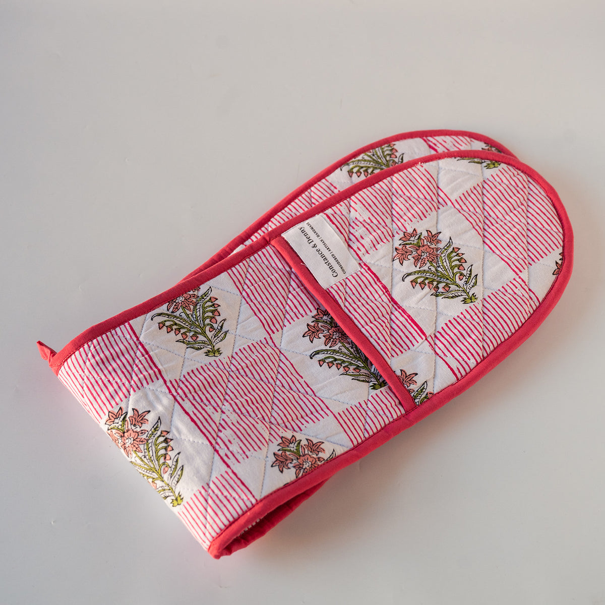 Two quilted cotton oven mitts with a PINK  and white striped and floral block print pattern, featuring a solid blue trim and a white brand label.