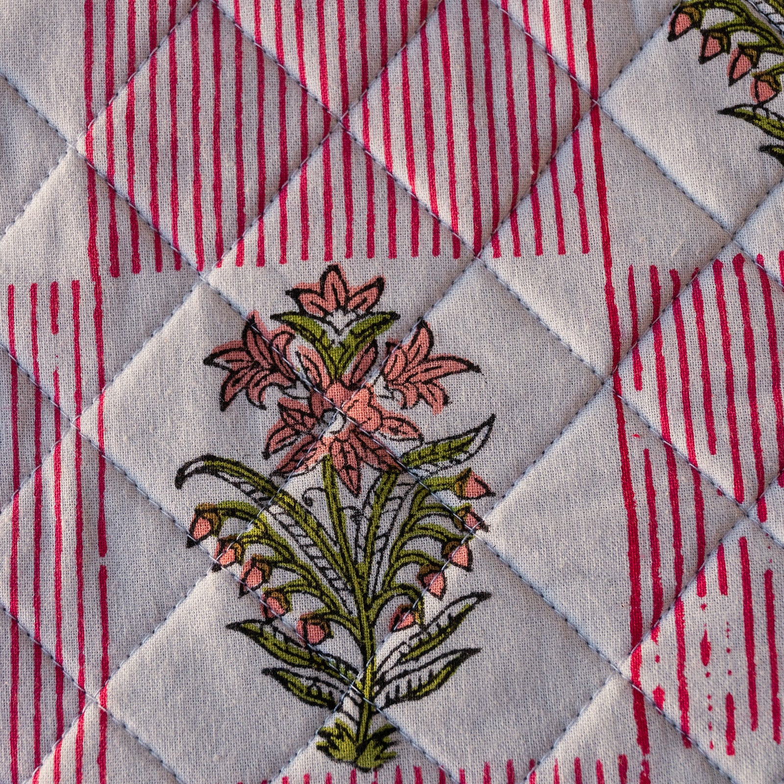 Close-up of a quilted textile with a diamond-stitched pattern, pink and red vertical stripes, and embroidered flowers in pink, green, and white.