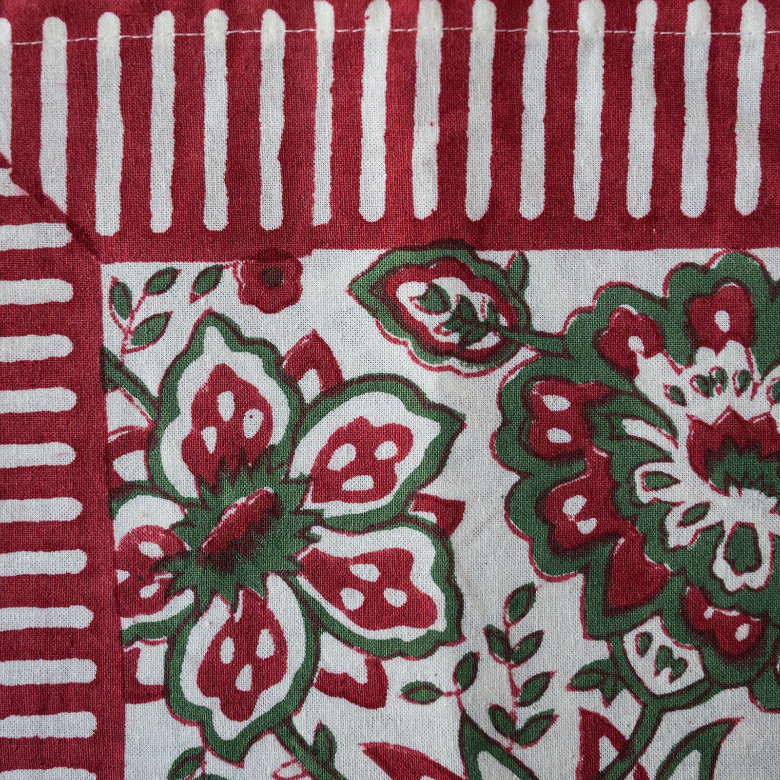 A close-up of a textile or fabric pattern with a red and white striped border at the top and left, and a central floral block-print style design in red, green, and white on an off-white background.