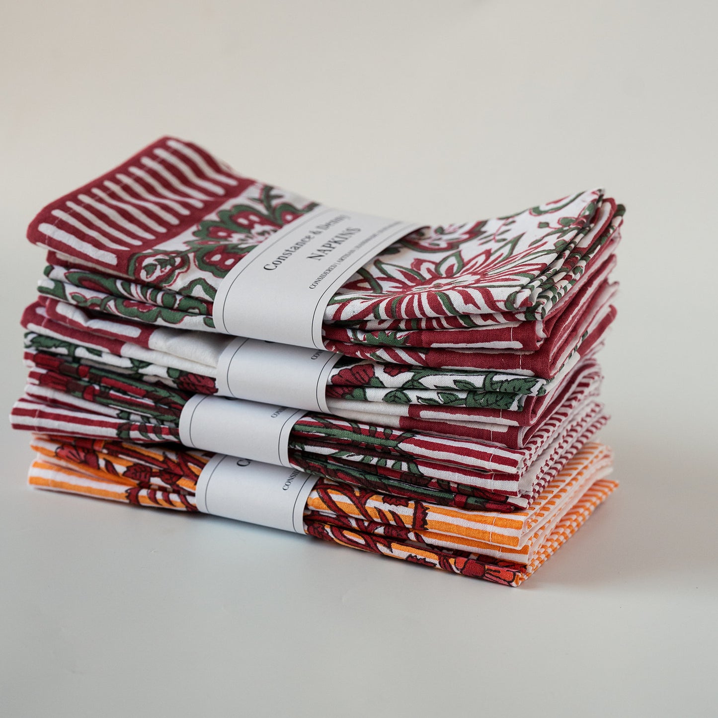 A stack of folded cotton kitchen towels with various holiday and autumn patterns, including red and white stripes, red and green floral motifs, and orange and brown leaf designs, wrapped with white paper bands.