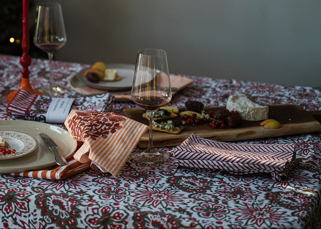 A cozy holiday table setting with a red and white patterned tablecloth, lit candles in red and orange holders, wine glasses, and charcuterie boards with cheese and crackers. A blurred Christmas tree with warm lights is in the background.