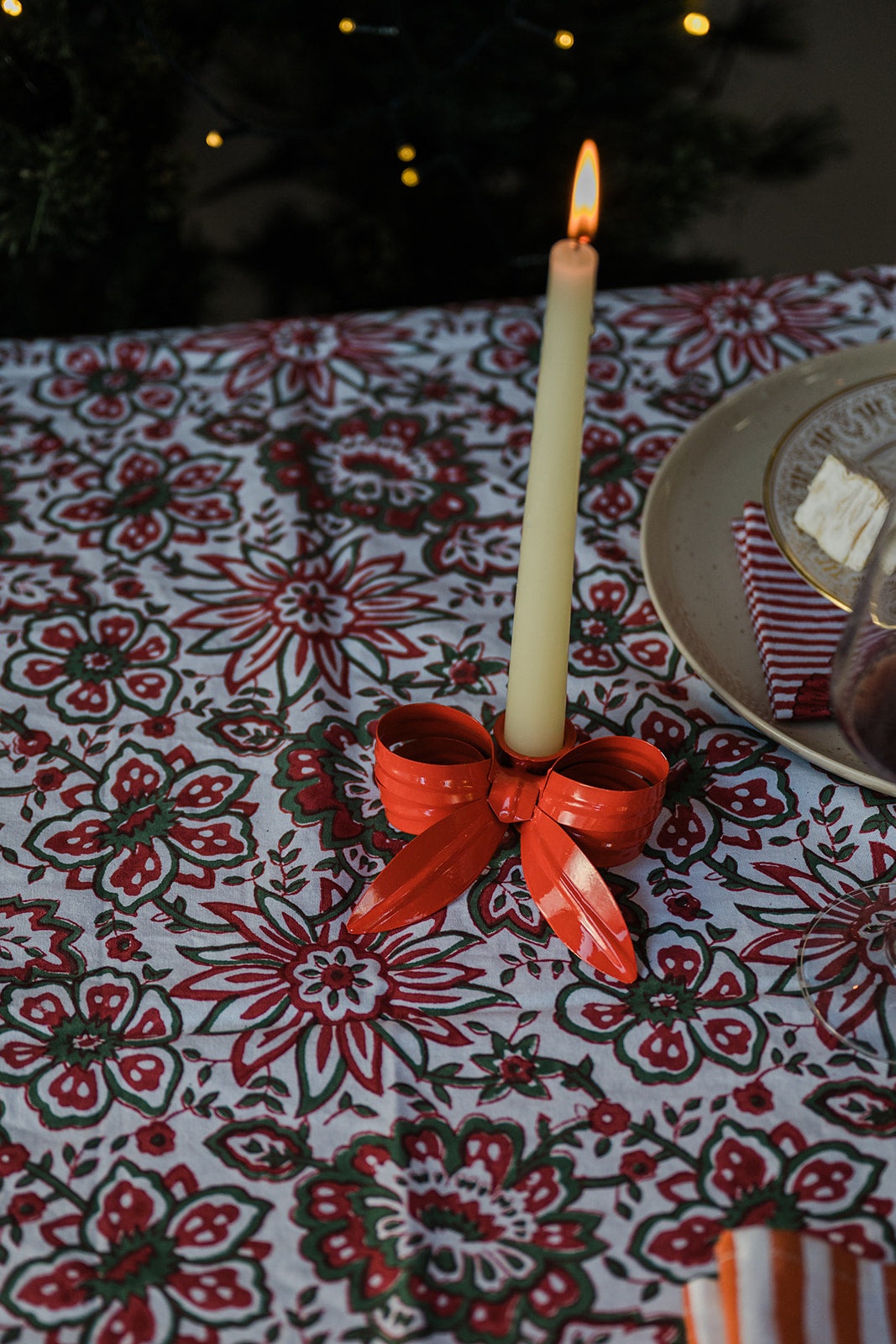 A lit cream taper candle in a red patterned holder with a matching red bow, set on a red and white floral tablecloth next to a plate.