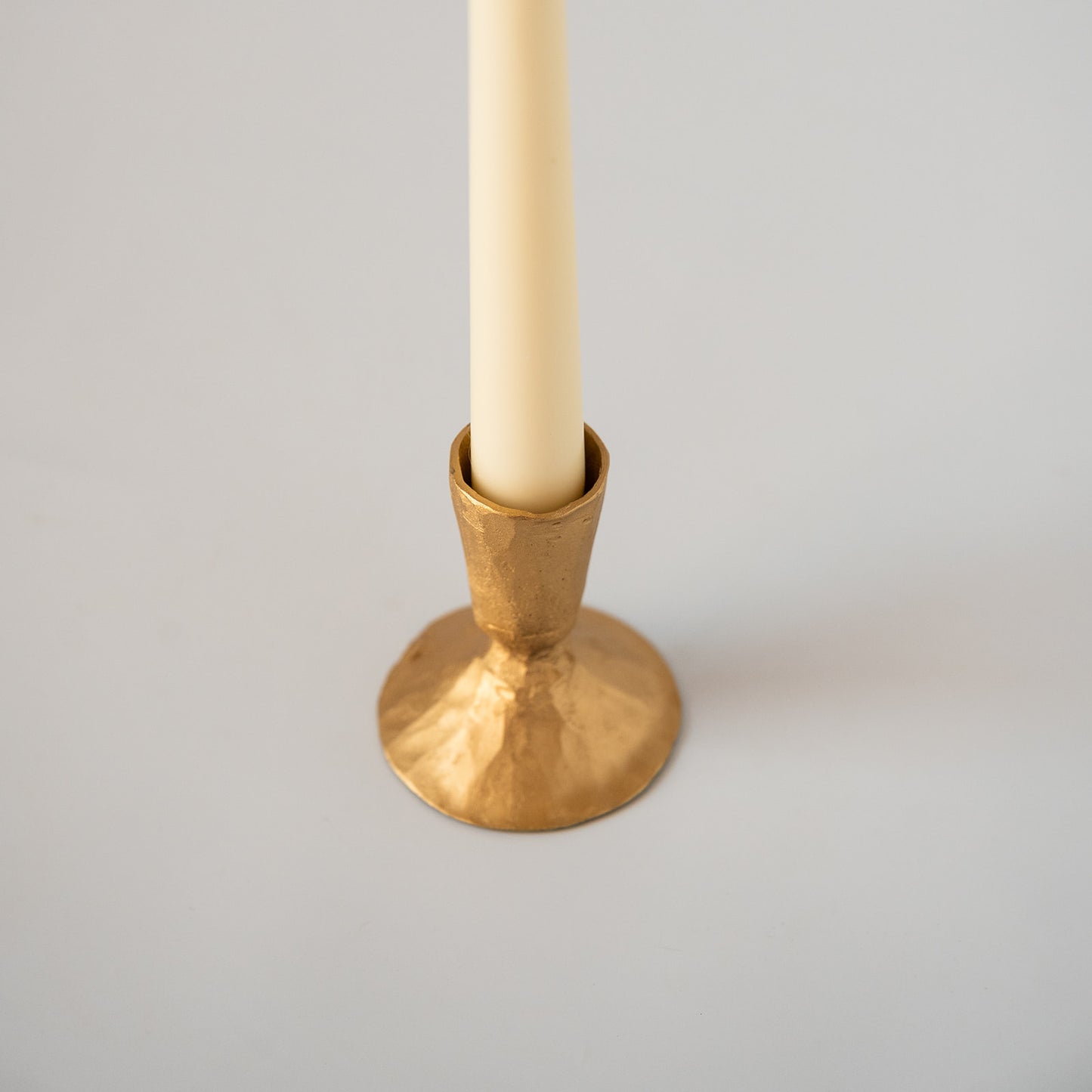 A hammered gold brass candle holder with a textured finish, holding a cream-colored taper candle, set against a neutral light gray background.