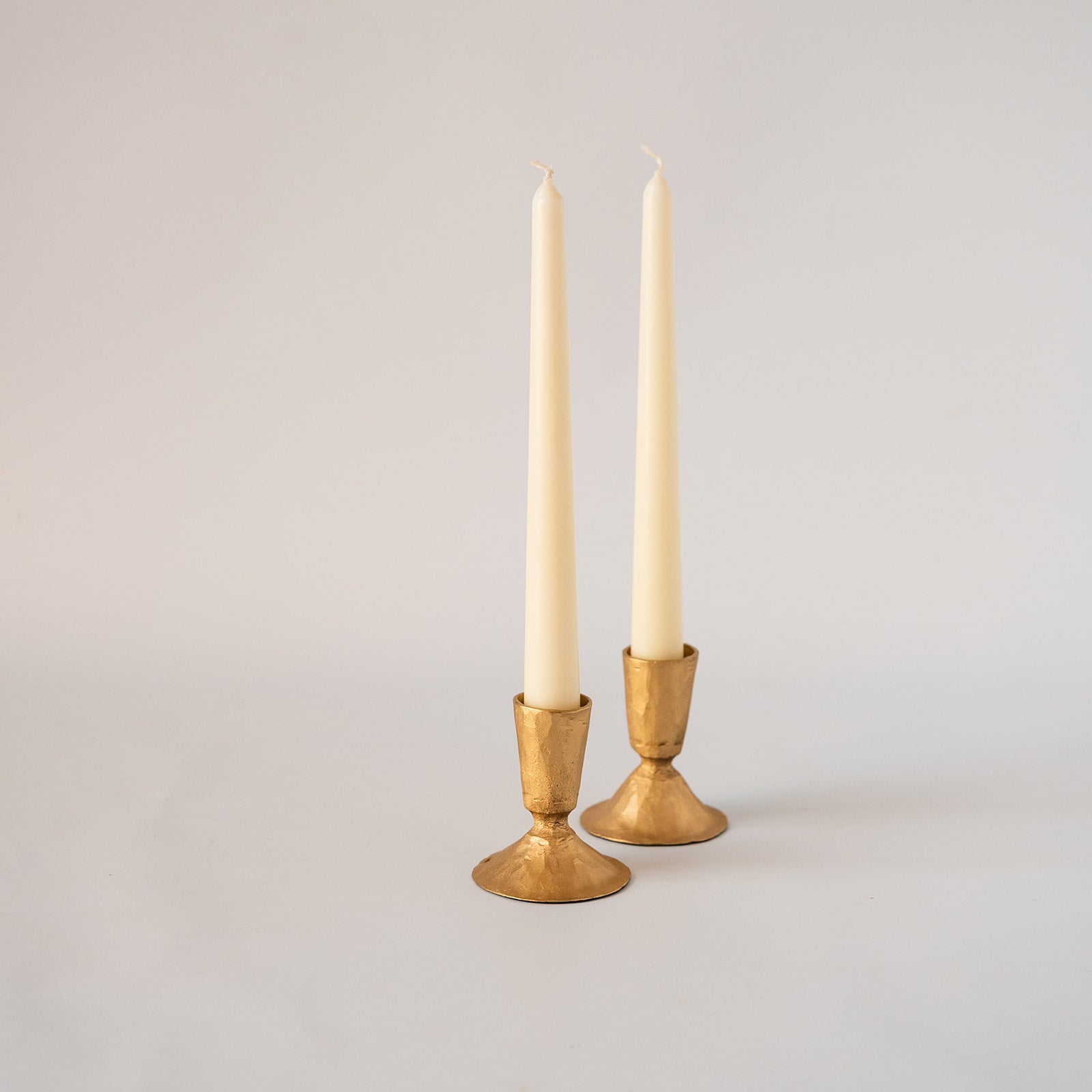 Two cream taper candles in hammered gold metal candlesticks, photographed against a seamless light gray studio background.