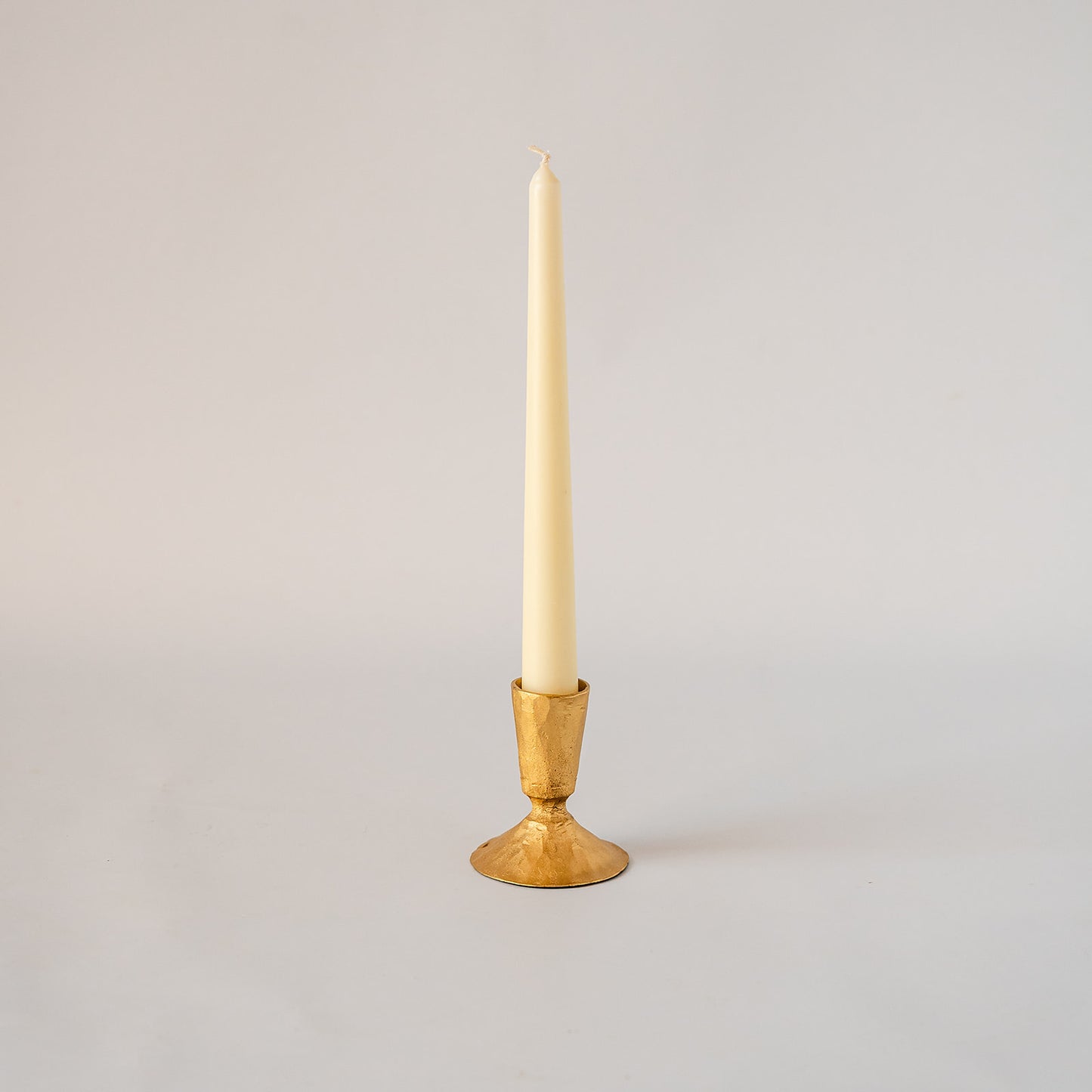 A single cream taper candle standing upright in a polished gold metal candlestick holder against a plain light gray studio background.