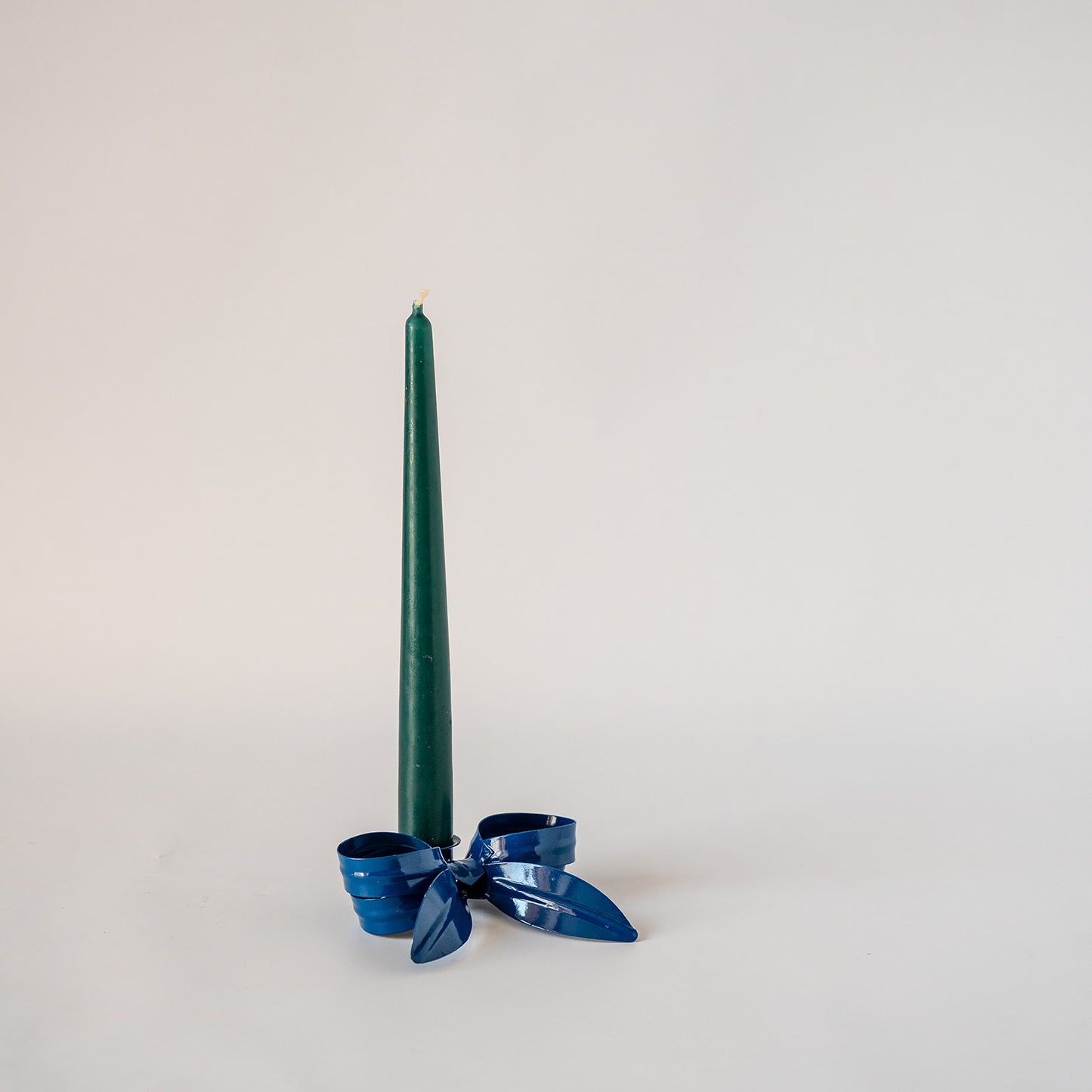 A dark blue taper candle standing upright in a shiny blue metal candlestick holder with a decorative bow, set against a plain light gray background.