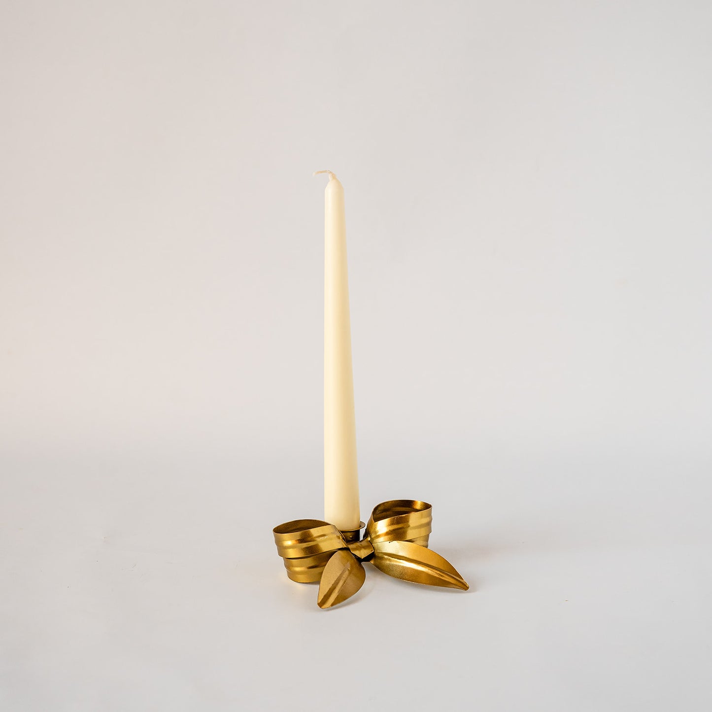 A single cream taper candle standing upright in a shiny gold metal holder shaped like a bow, photographed against a plain light gray studio background.