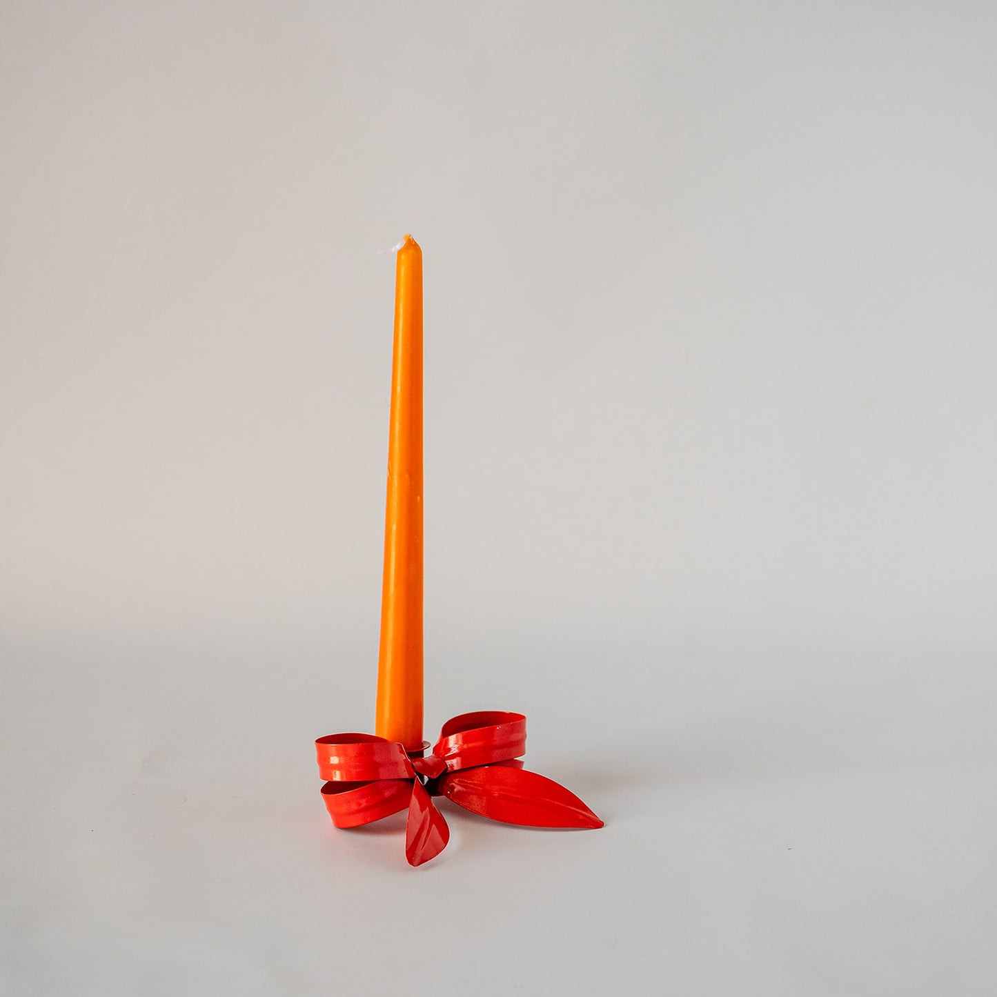 A single orange taper candle standing upright in a red ribbon holder against a plain light gray background.