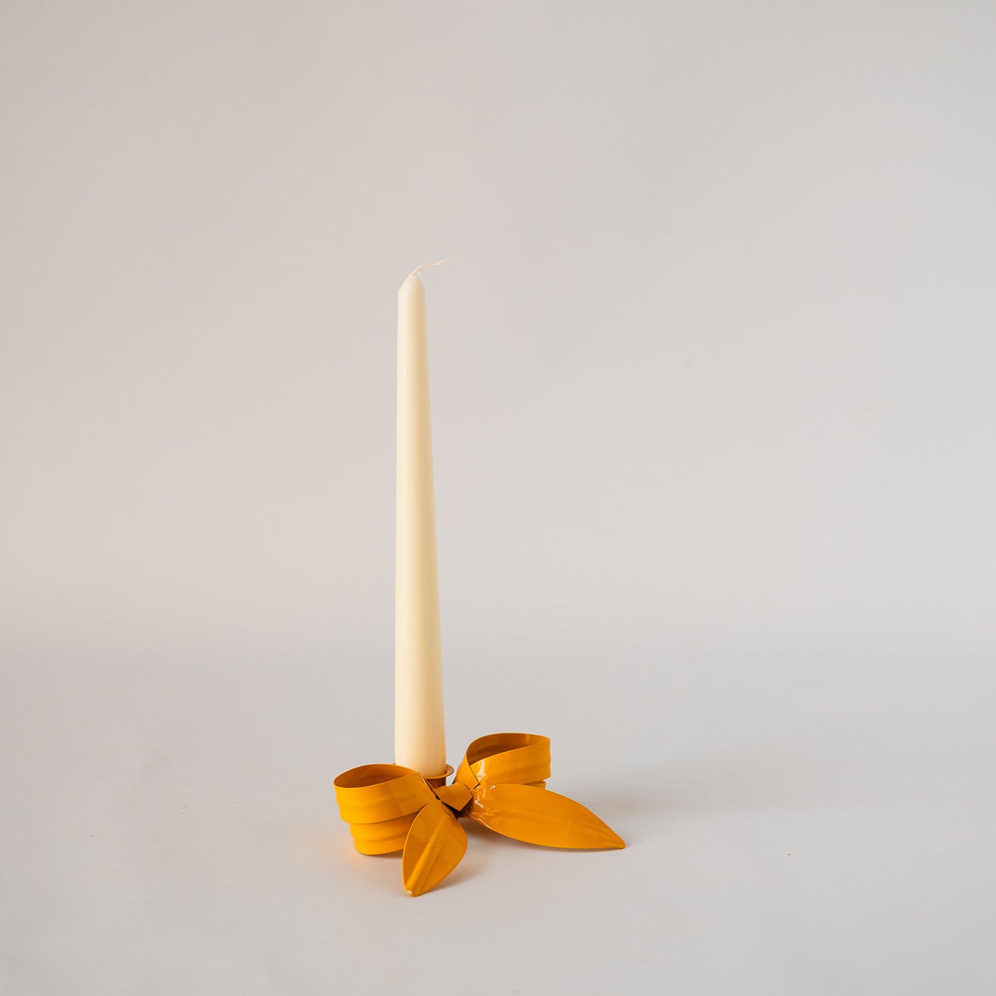 A single cream taper candle standing upright in a small yellow ribbon holder with a bow, photographed on a plain light gray studio background.