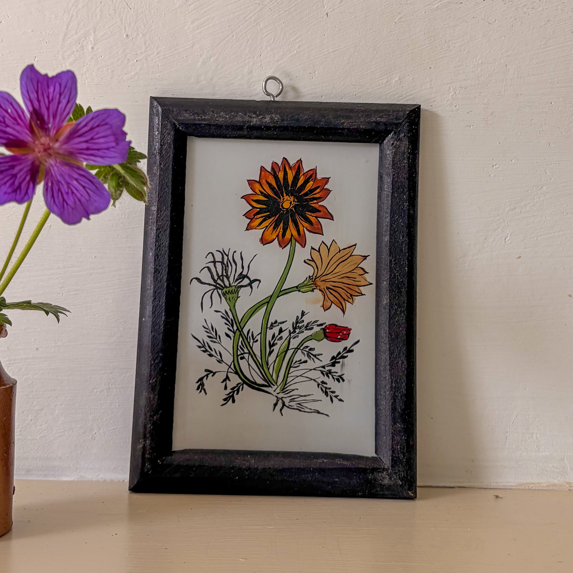 Framed artwork of flowers on a white wall with a purple flower in the foreground.