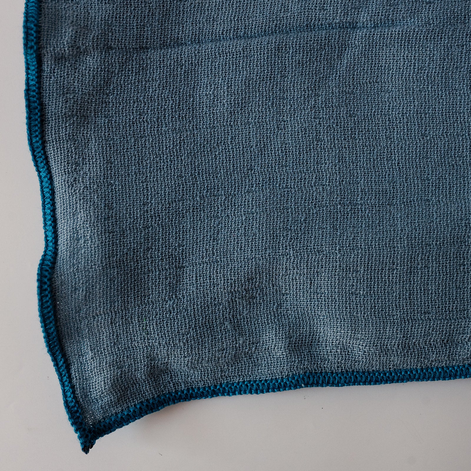 A close-up of a dark teal napkin showing its textured terry fabric and matching braided edge on a light gray background.
