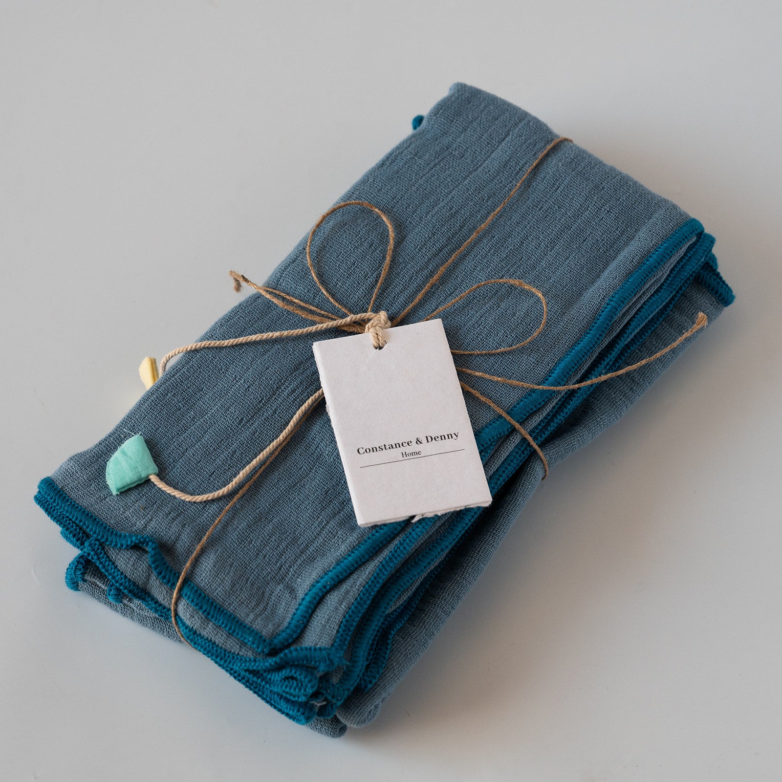 A folded stack of textured blue and teal fabric napkins tied with twine and a paper tag reading 'Constance & Dream', on a light gray background.